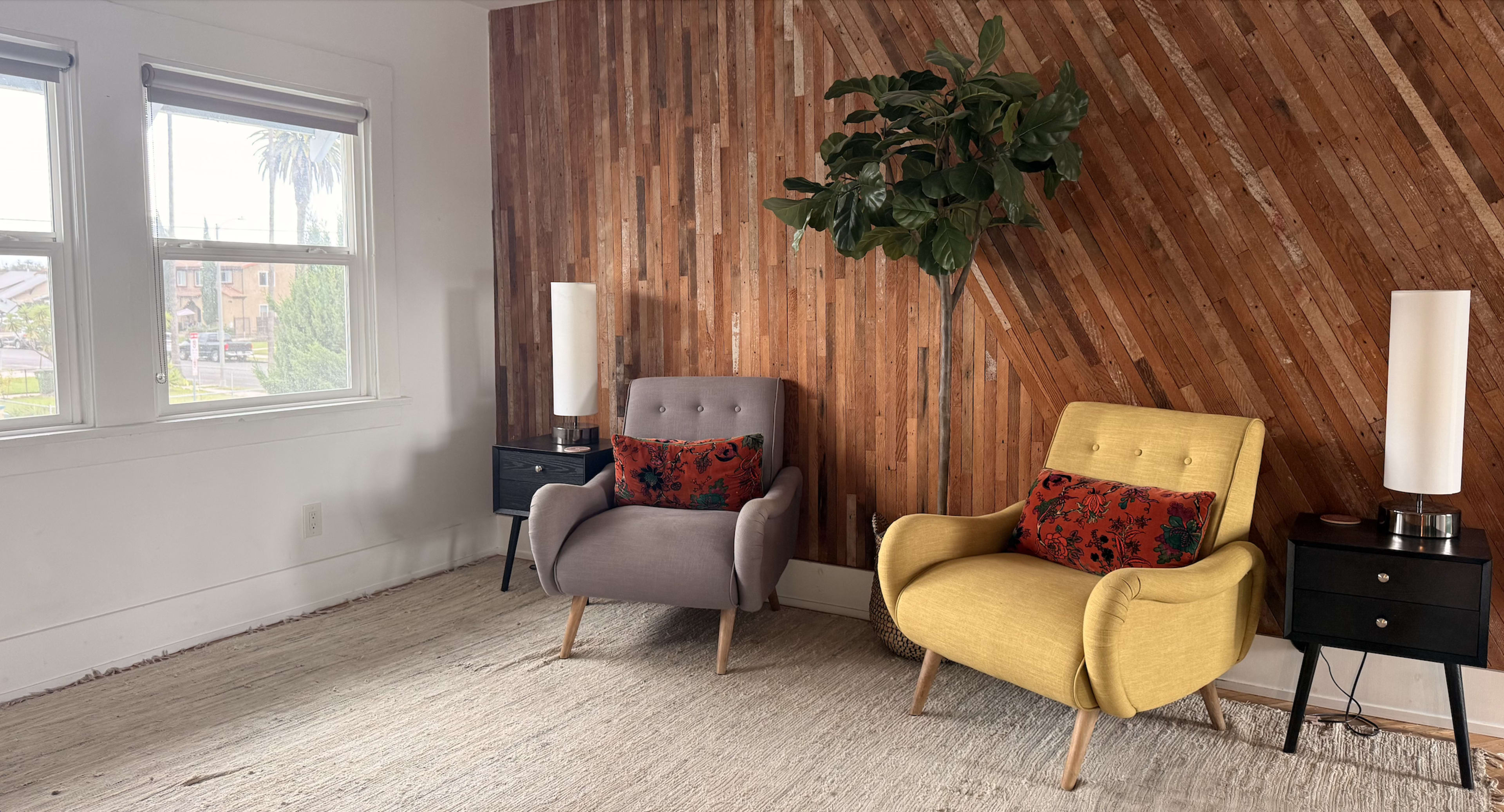 The image shows a room with two vintage-style armchairs, one gray and one yellow, placed on a light-colored rug against a wooden feature wall, with side tables and lamps on either side.