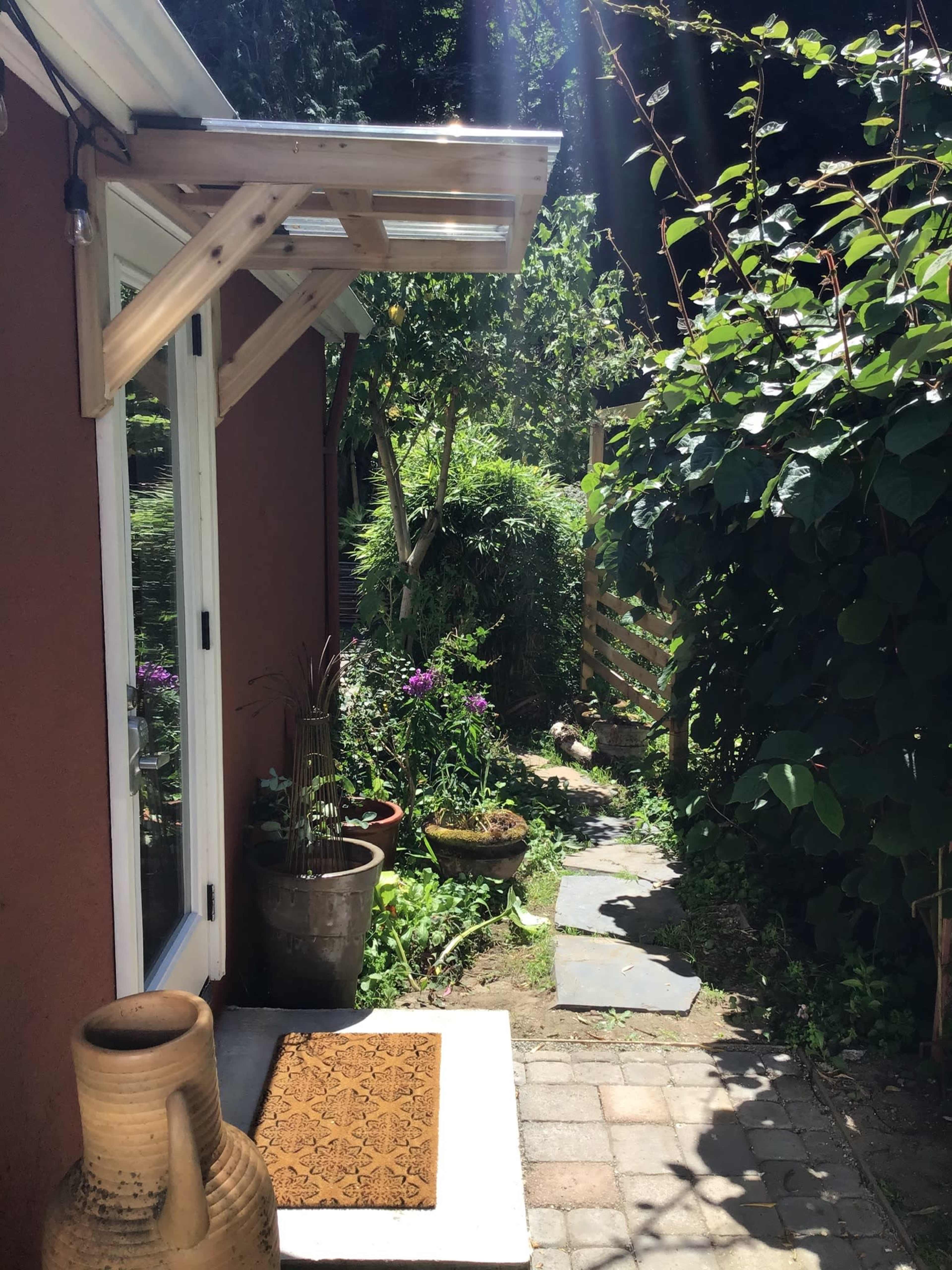 A stone pathway leads through a garden with potted plants and greenery beside a brown-walled house.