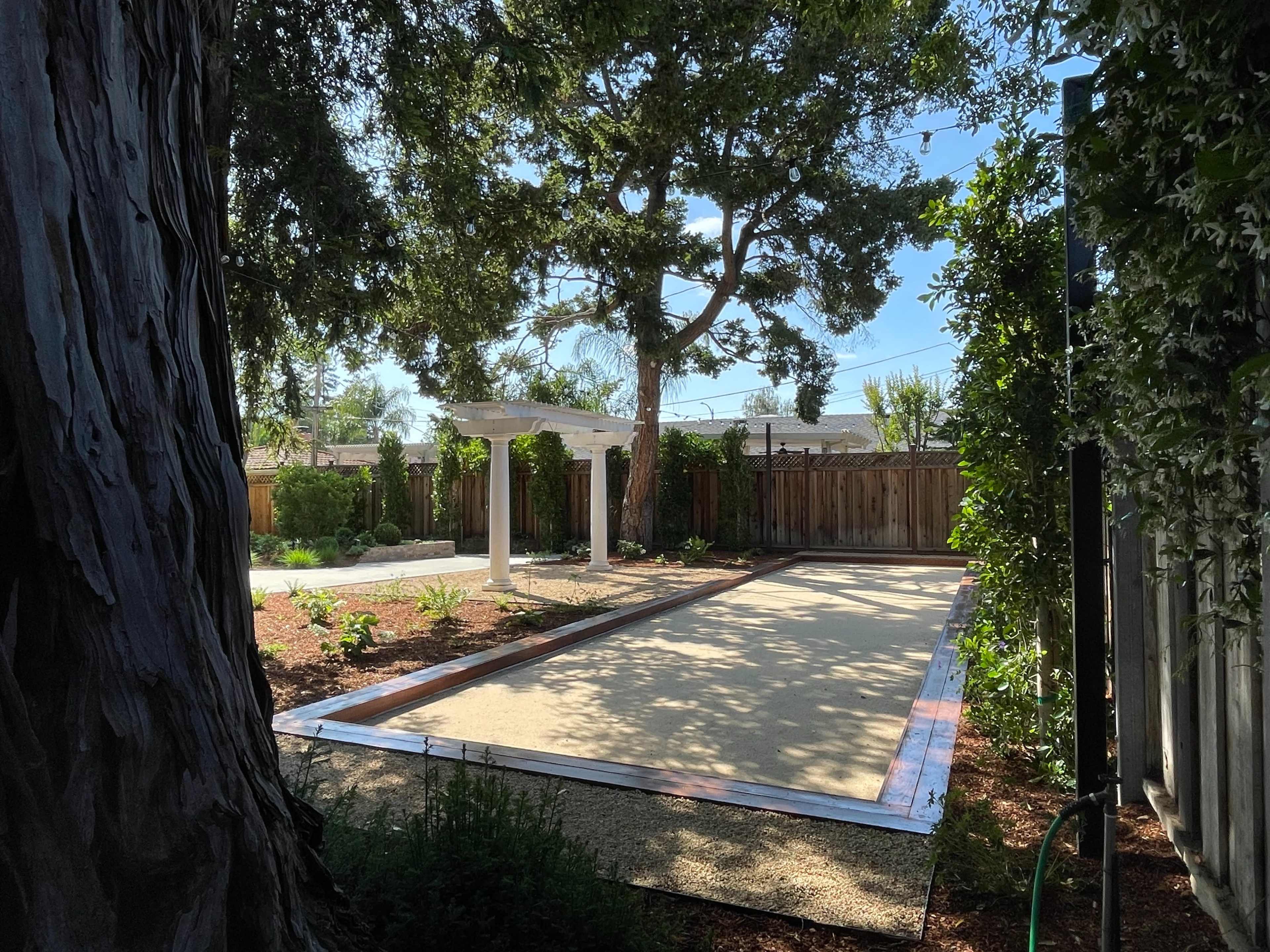 A sandy bocce ball court is set in a landscaped backyard surrounded by greenery and wooden fencing.