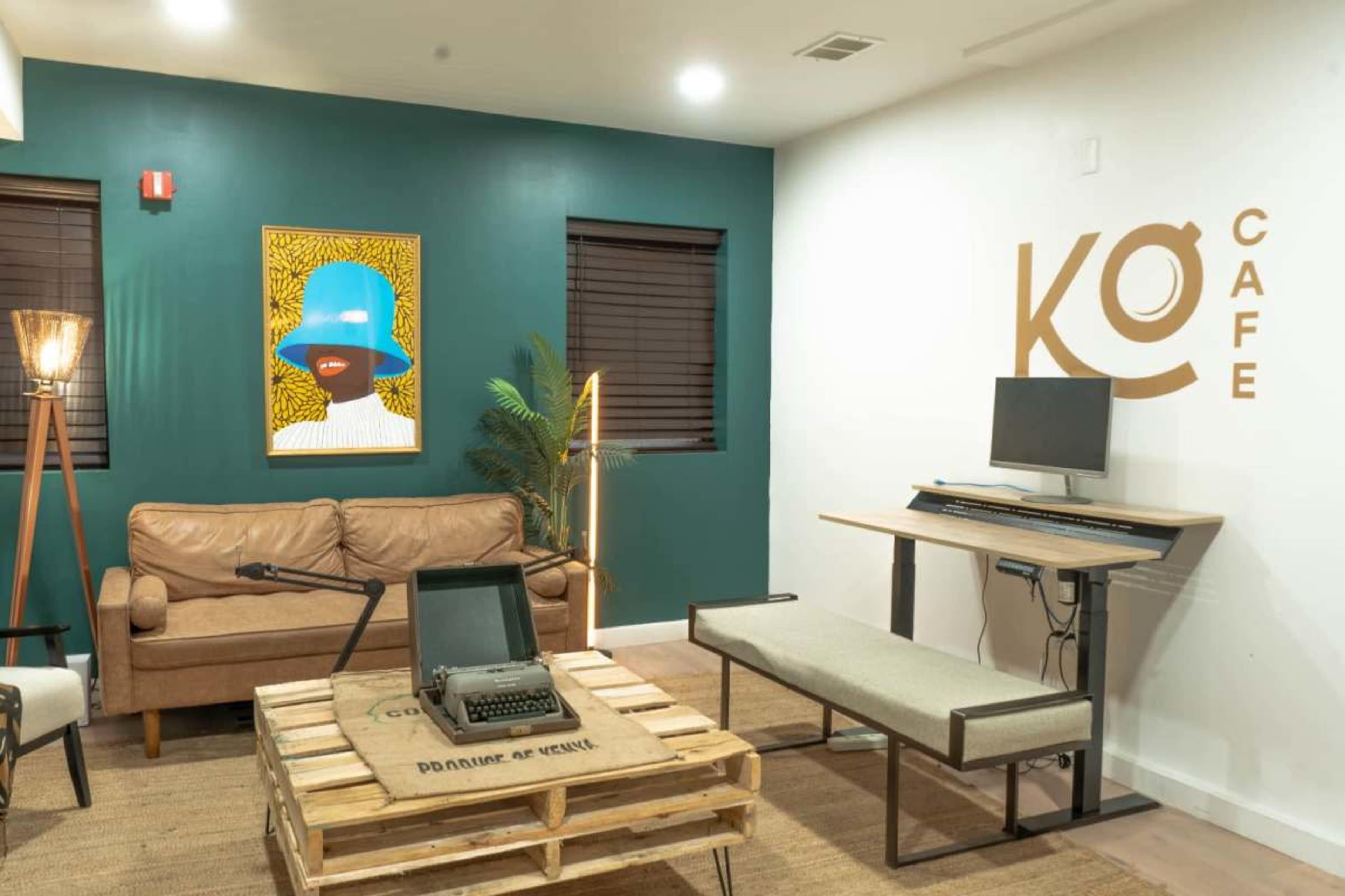 The image depicts a cozy cafe interior featuring a brown leather couch, a wooden pallet coffee table, an old typewriter, modern furniture, and a vibrant piece of artwork on the wall.