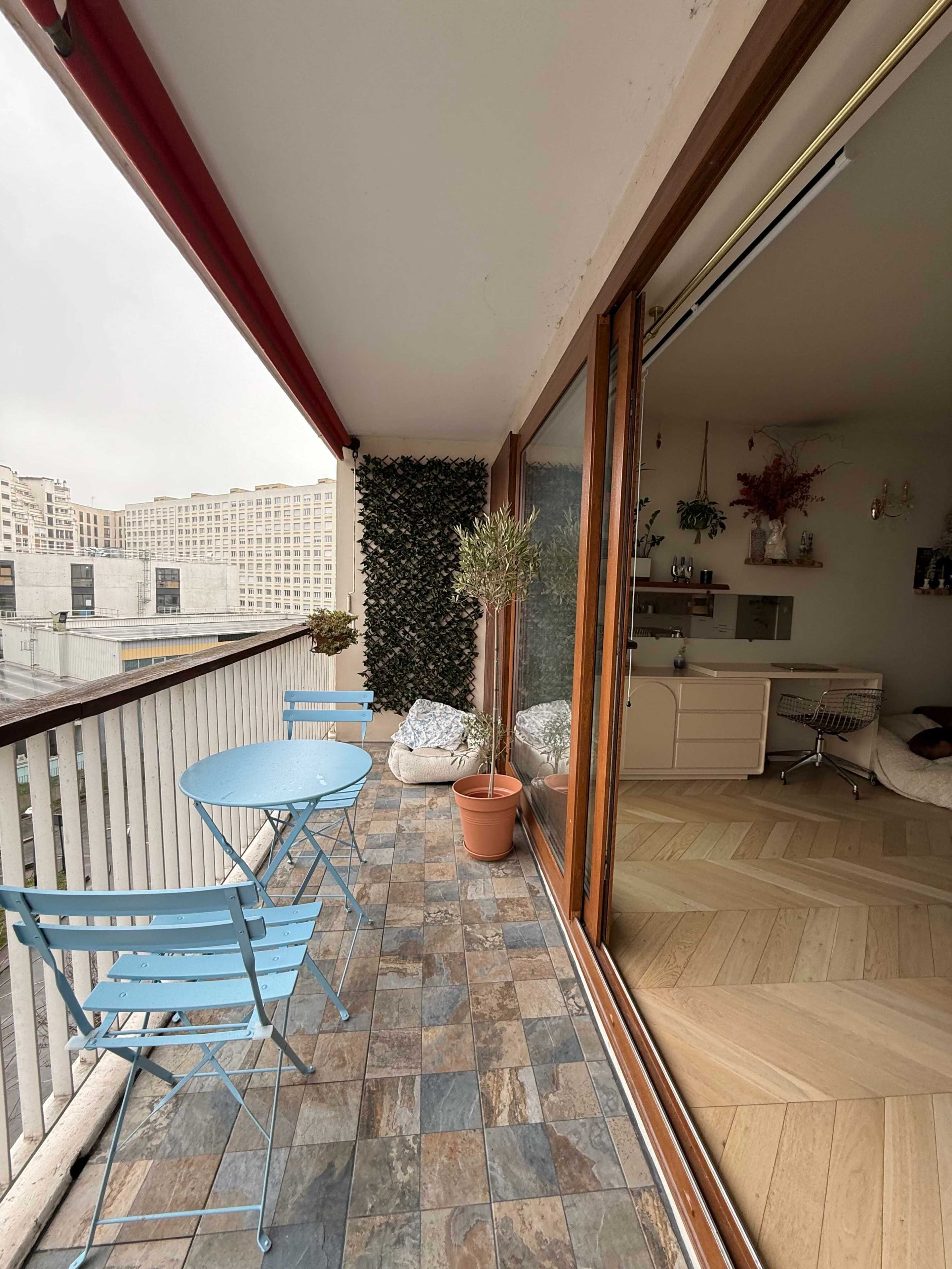 A balcony features a small blue table and chairs, potted plants, and a view of a cityscape through large glass doors.