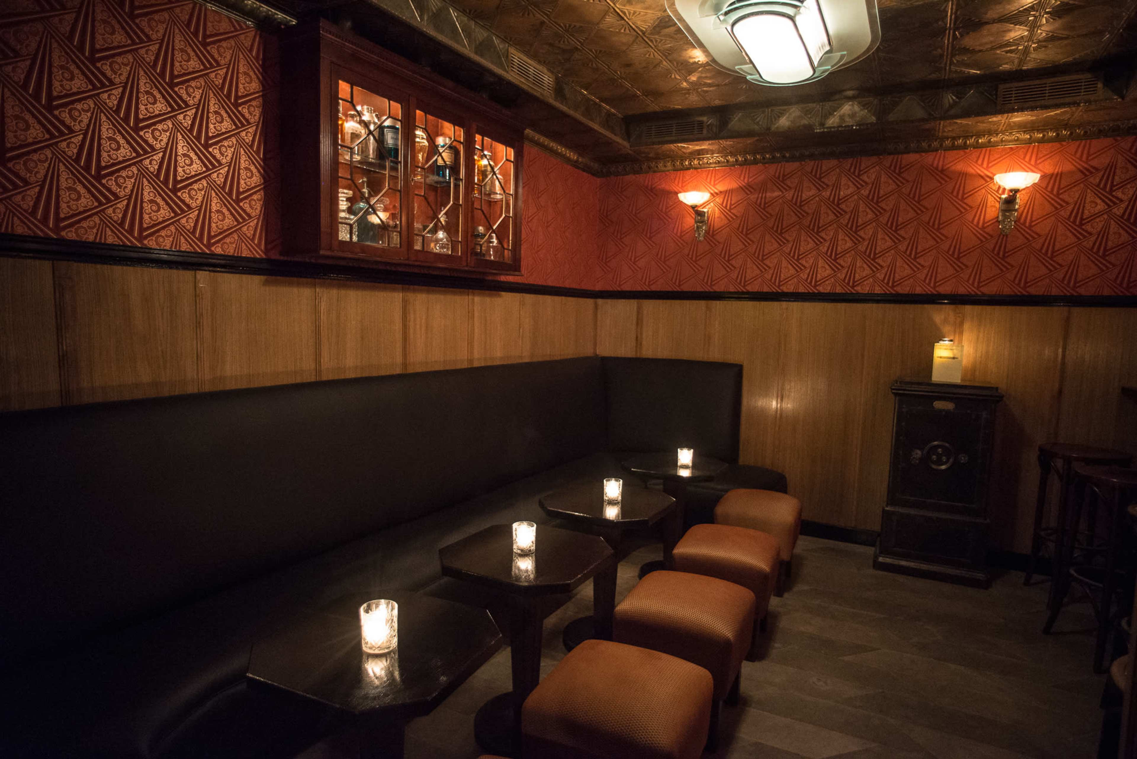 The image shows a dimly lit bar area featuring a cushioned seating arrangement with candlelit tables against a vintage-styled wall.