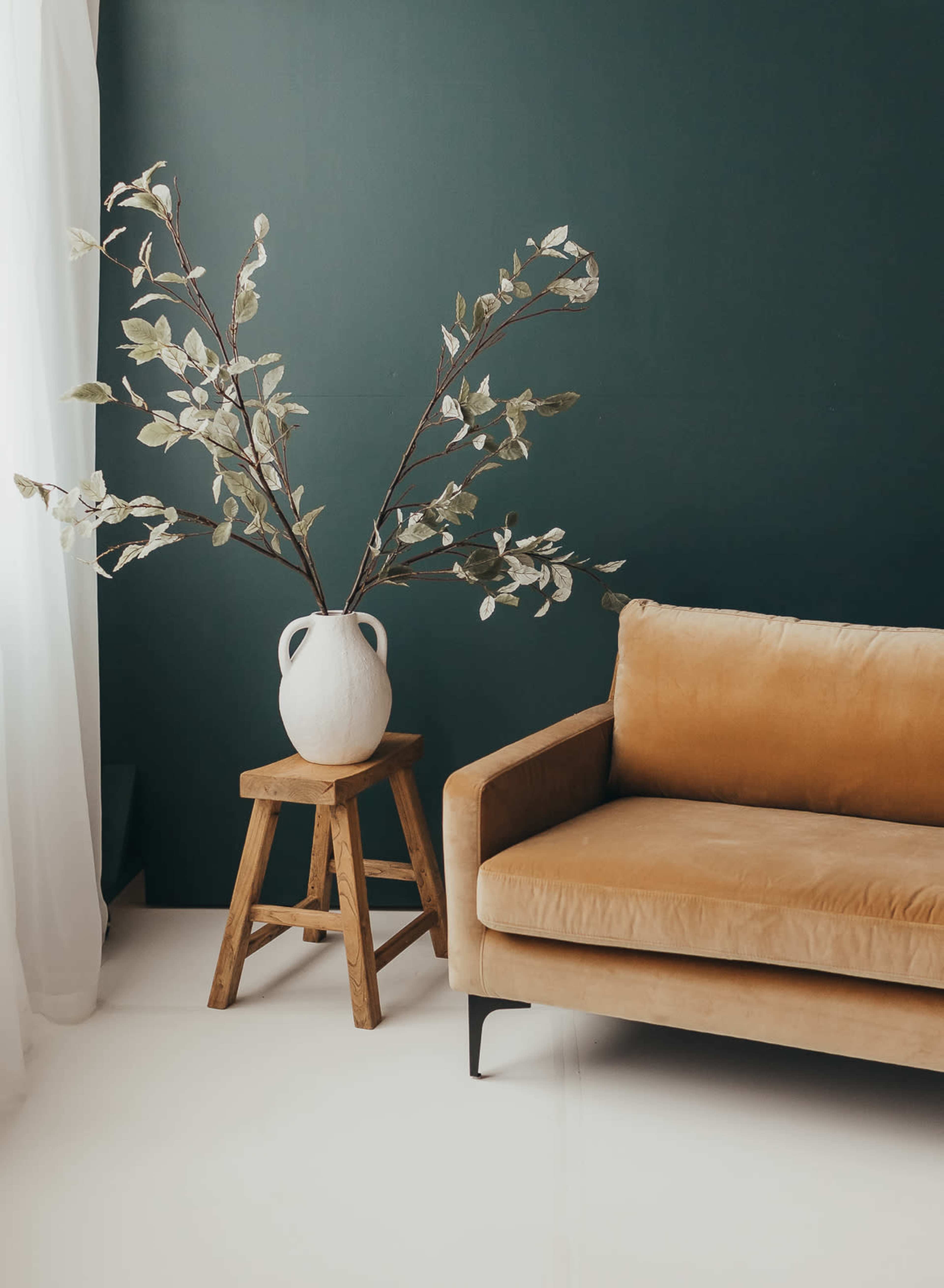 A brown velvet sofa is positioned next to a wooden stool holding a large white vase with branches against a dark green wall.