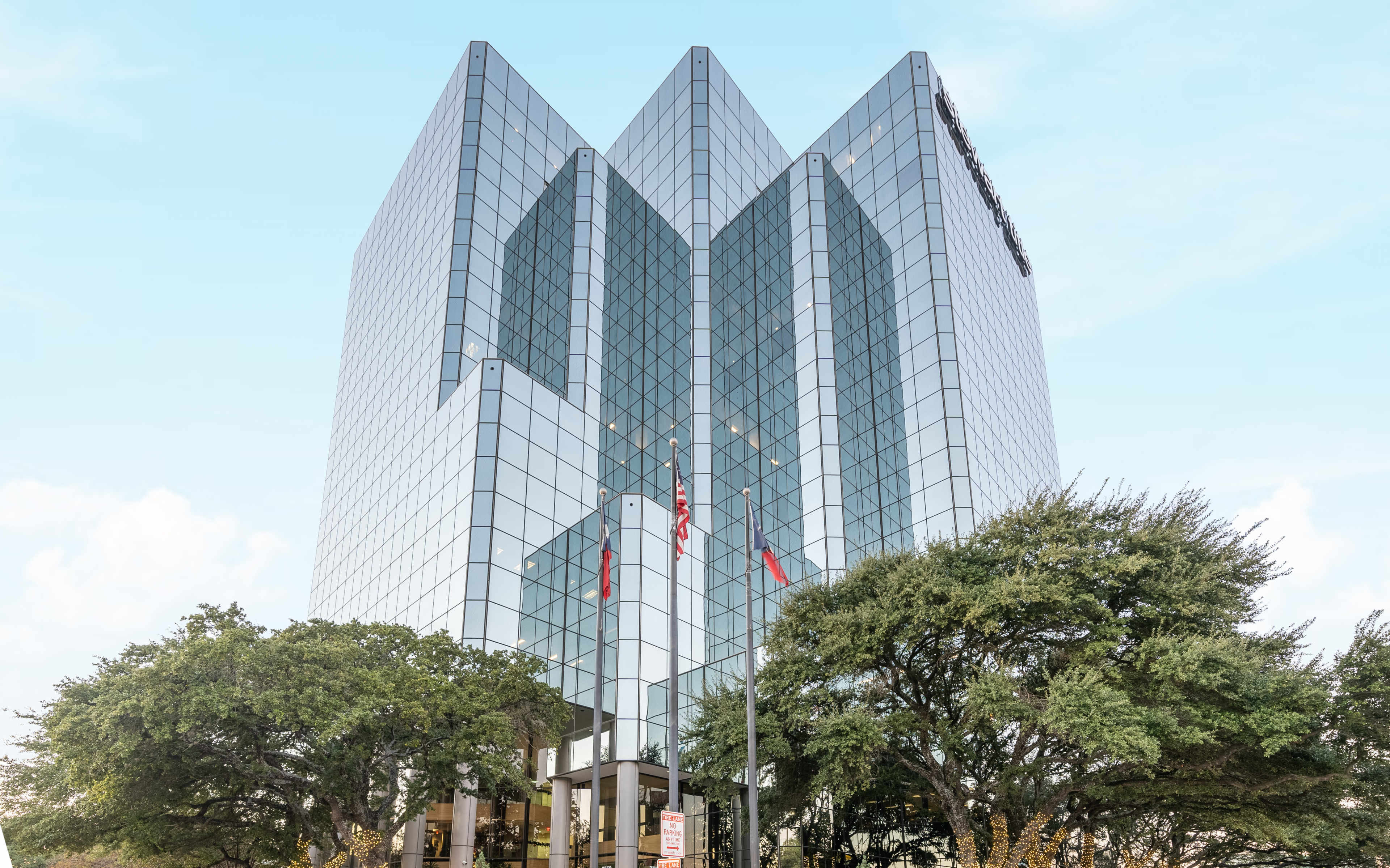 A tall glass office building with a modern geometric design stands amid trees and flags in front of its entrance.