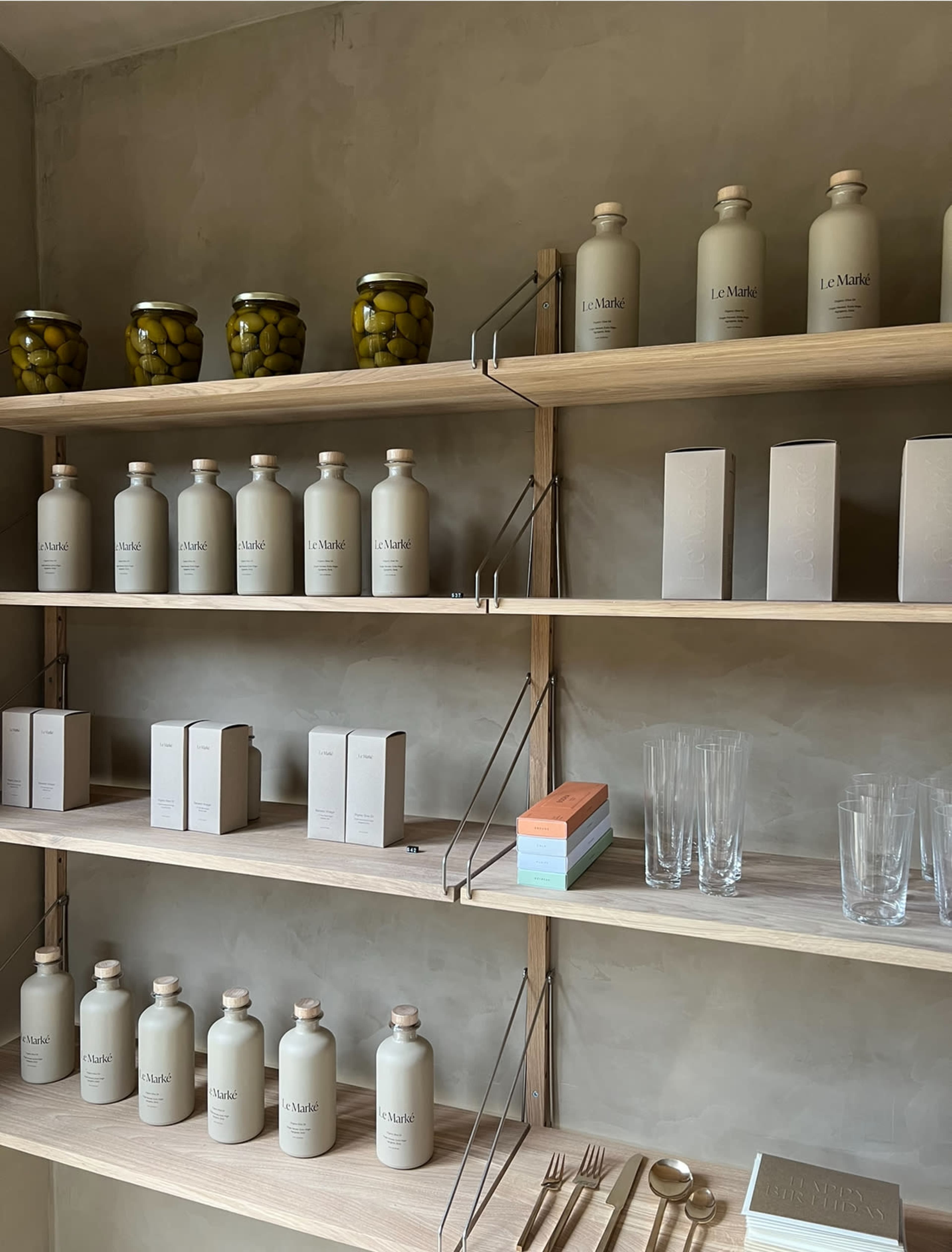 A wooden shelving unit displaying jars of olives, decorative bottles, boxed items, glassware, and utensils against a simple, muted wall.