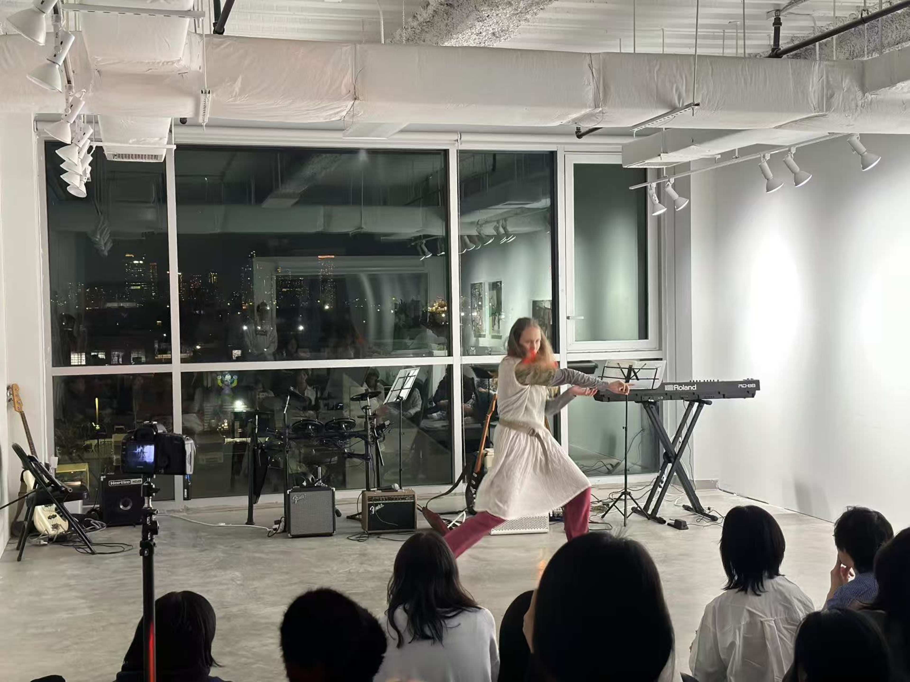 A performer in a white dress is dancing in front of an audience in a well-lit studio with large windows displaying a cityscape at night.