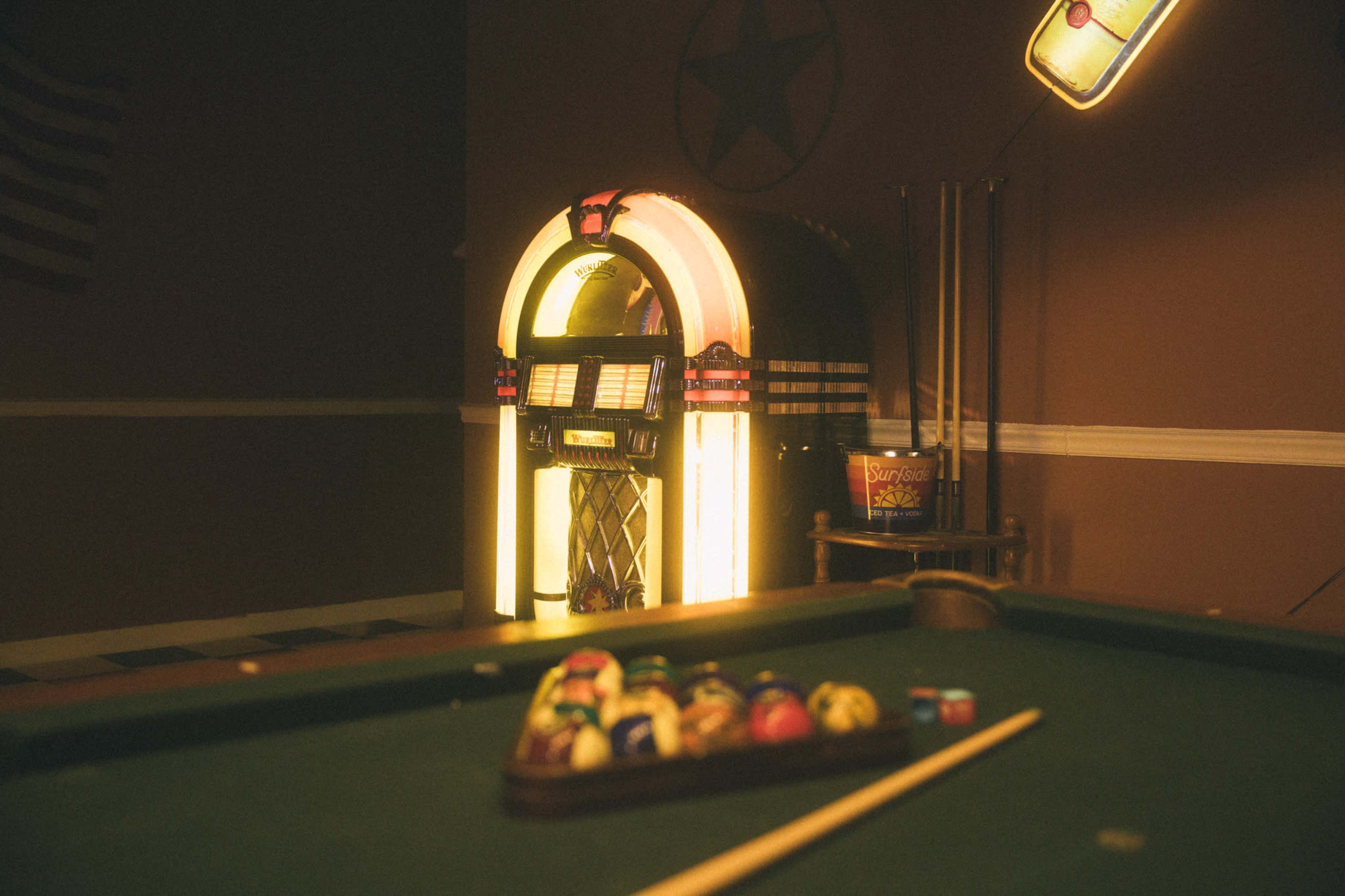 A vintage jukebox stands illuminated in a room with a pool table and a tray of billiard balls in the foreground.