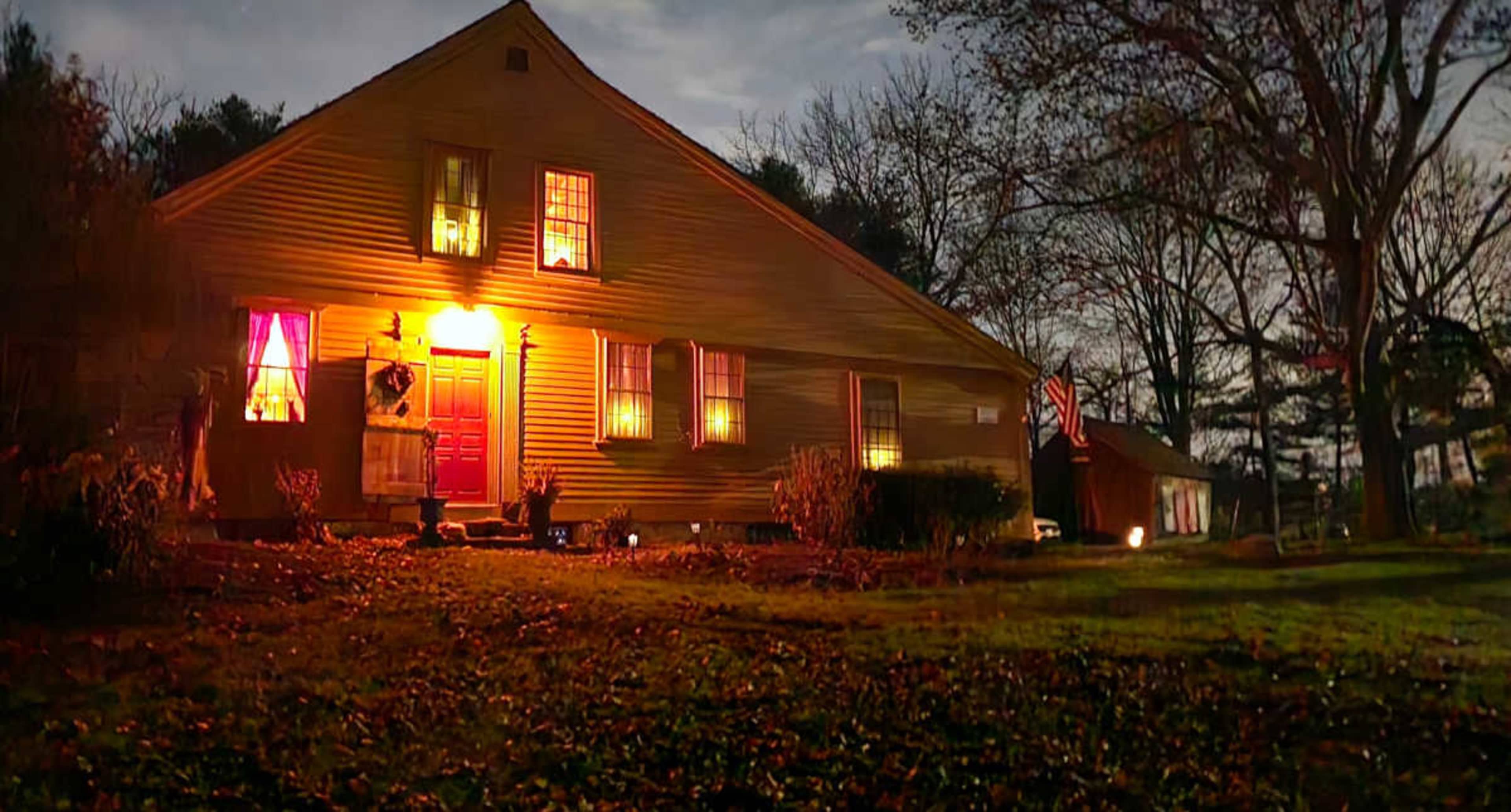 A yellow house with illuminated windows and a front door is shown at night, surrounded by trees and a grassy lawn.