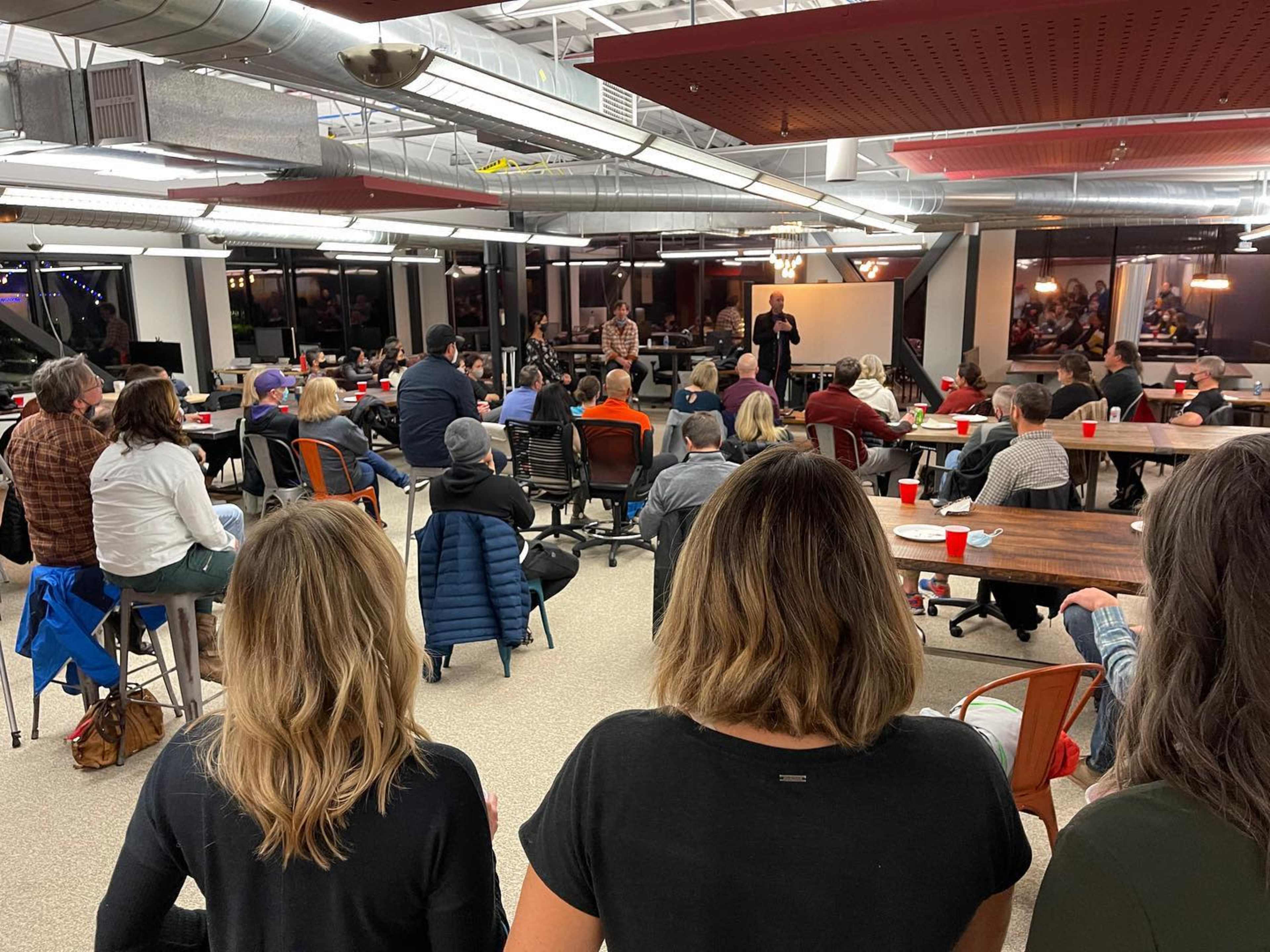 A group of people is seated in a large, open room, facing a speaker at the front, with tables and chairs arranged for an event.