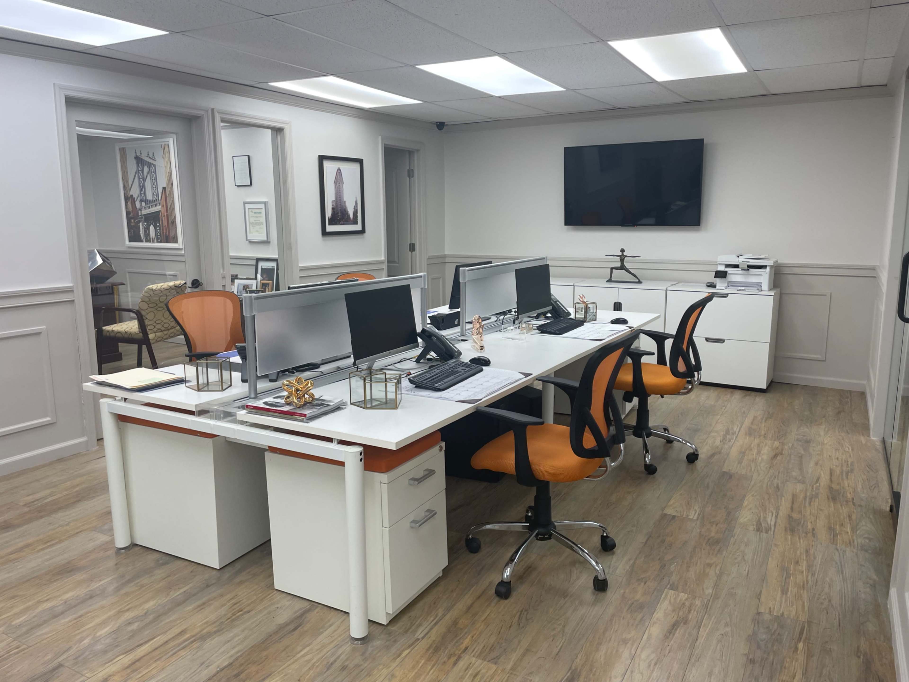 A modern office space with two white desks, each equipped with dual computer monitors, surrounded by orange office chairs, and a printer station in the background.