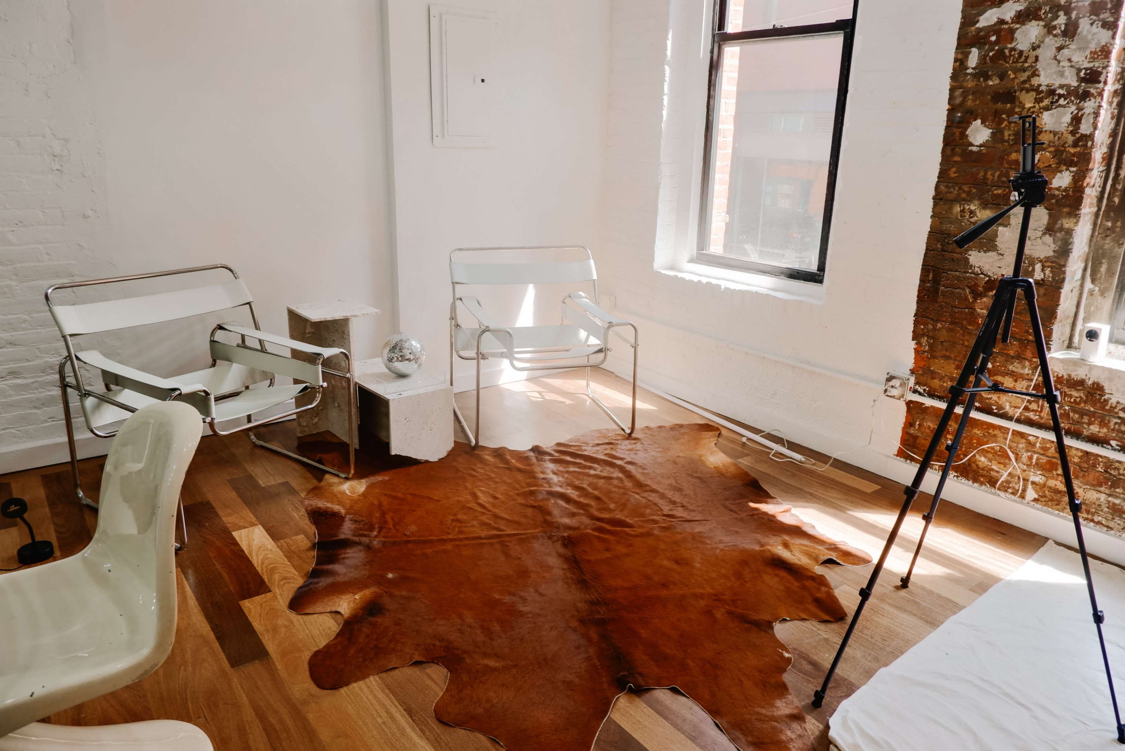 A minimalist room with two chairs, a tripod, a cowhide rug on the wooden floor, and a window revealing a brick wall.