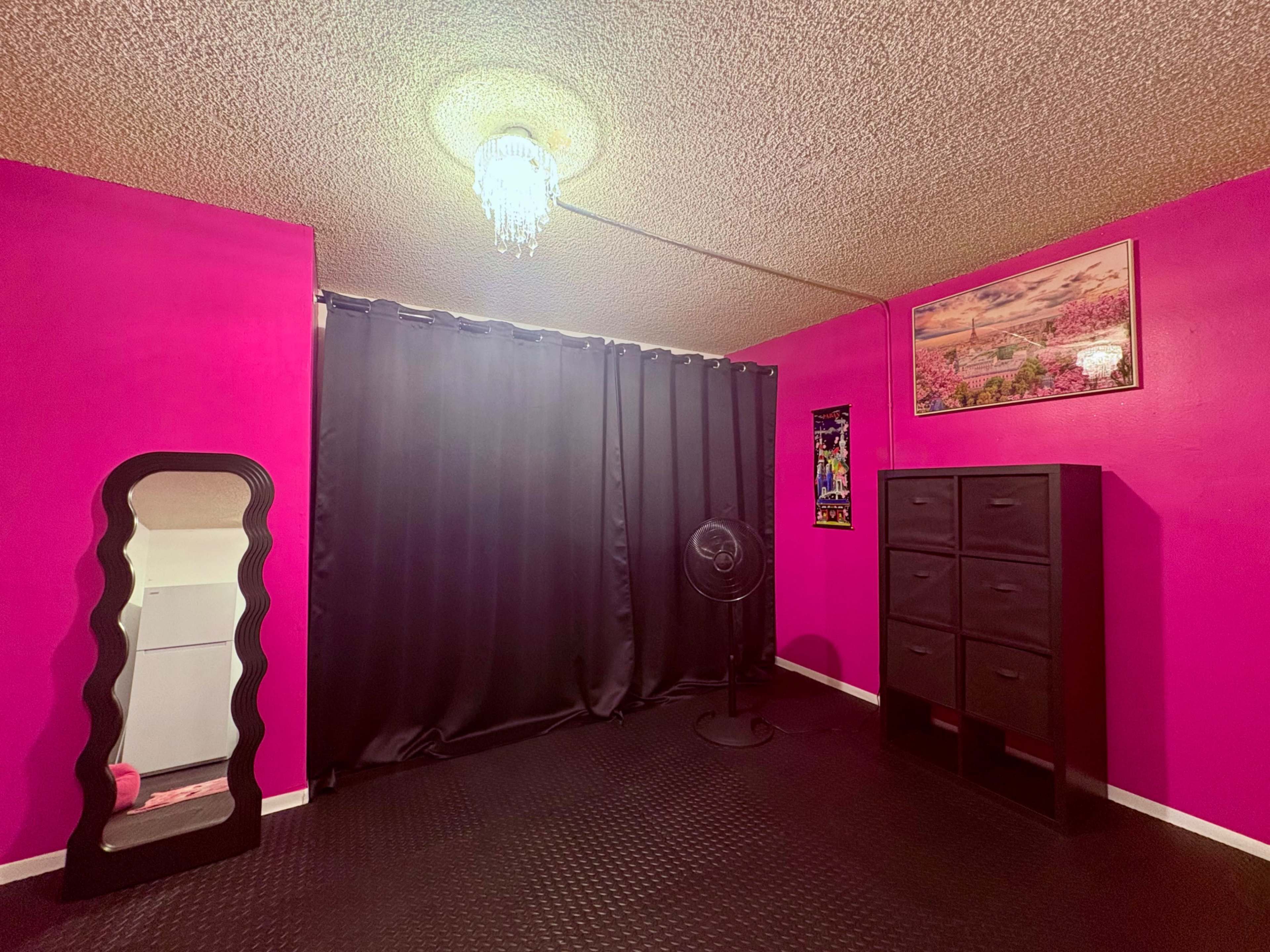 The image shows a room with bright pink walls, a black curtain, a mirror, a standing fan, and a black storage cabinet.