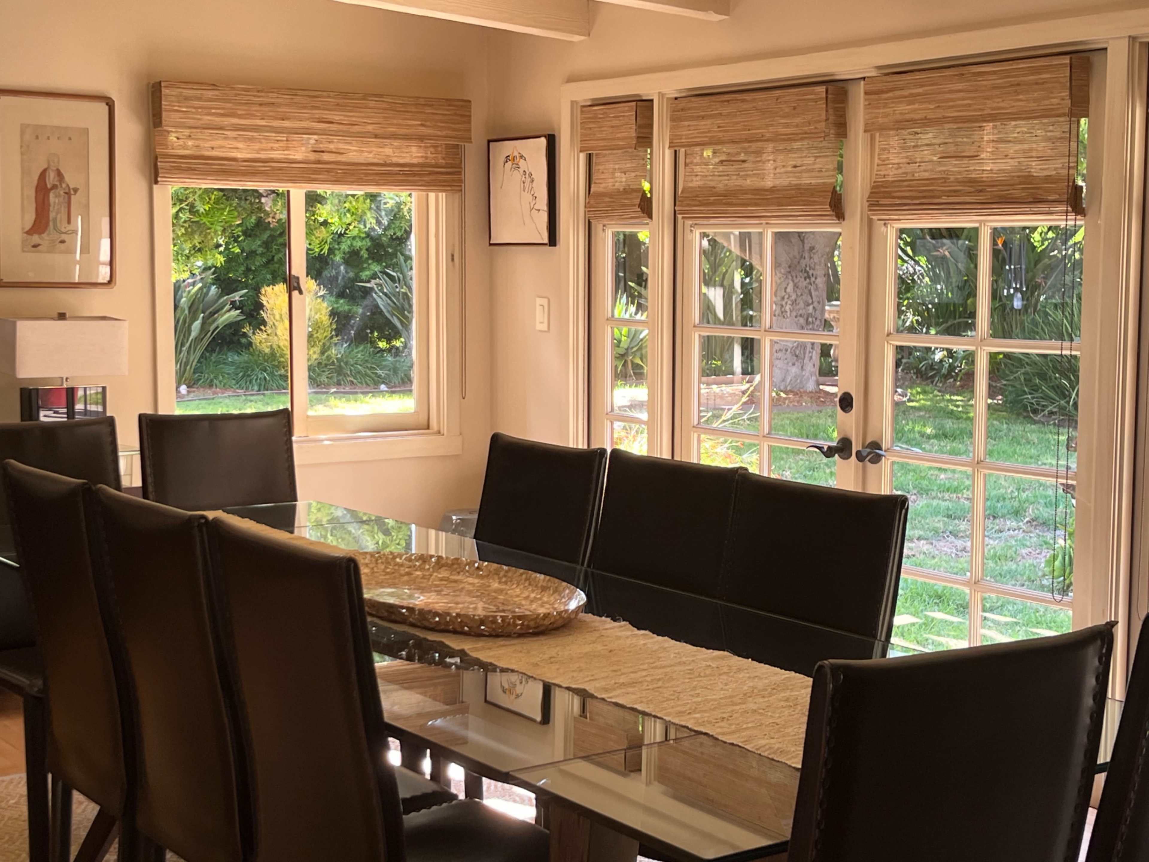 The image shows a dining room with a rectangular glass table surrounded by black chairs, illuminated by natural light coming through windows and doors that lead to a green outdoor space.