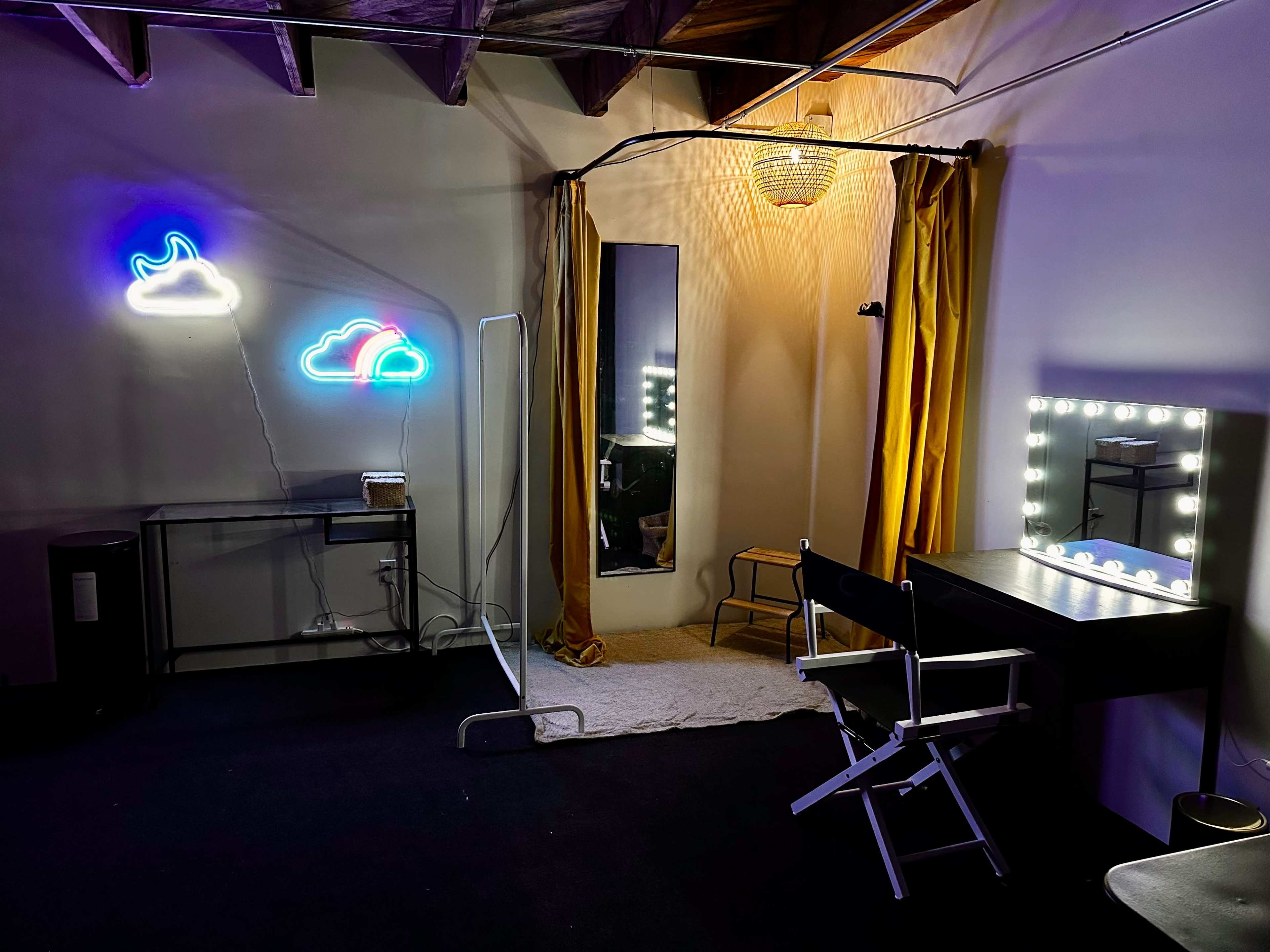 A dimly lit room featuring a makeup vanity with bright lights, a mirror, a curtain, and neon cloud decorations on the walls.