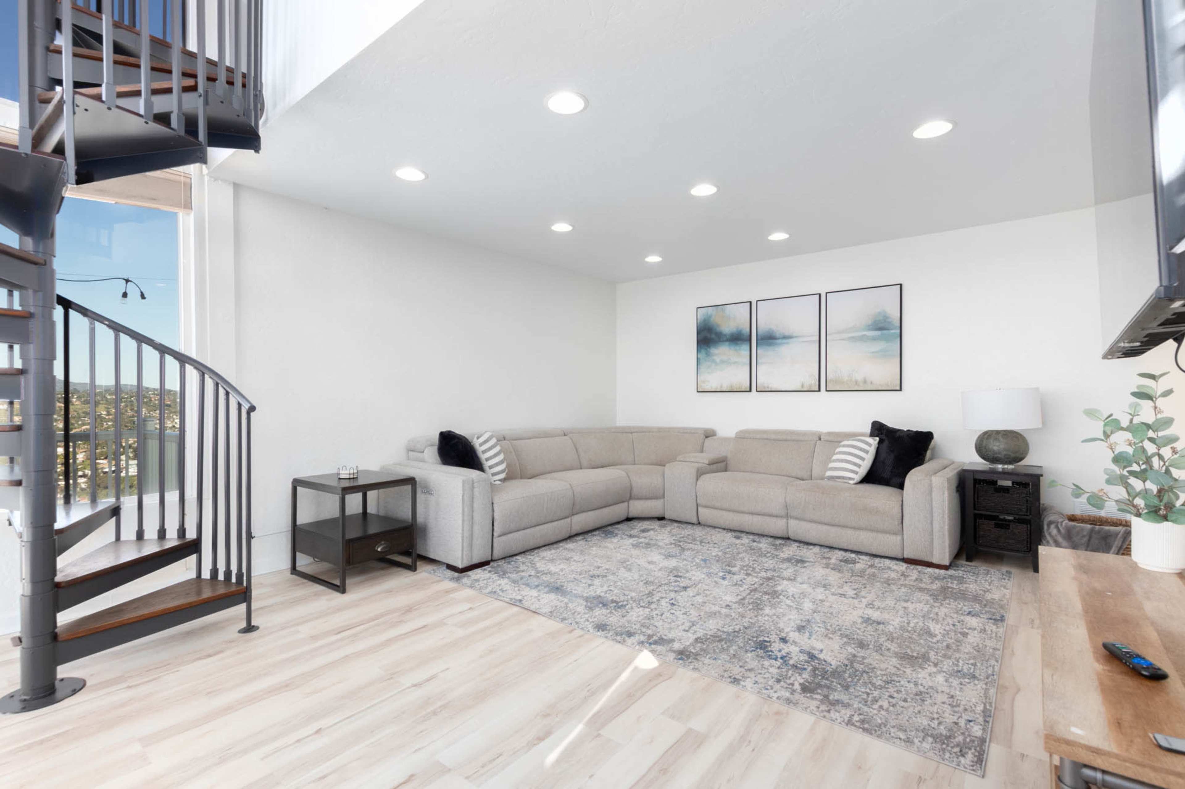 A modern living room features a gray sectional sofa, a spiral staircase, and wall art above a wooden table, with large windows allowing natural light.