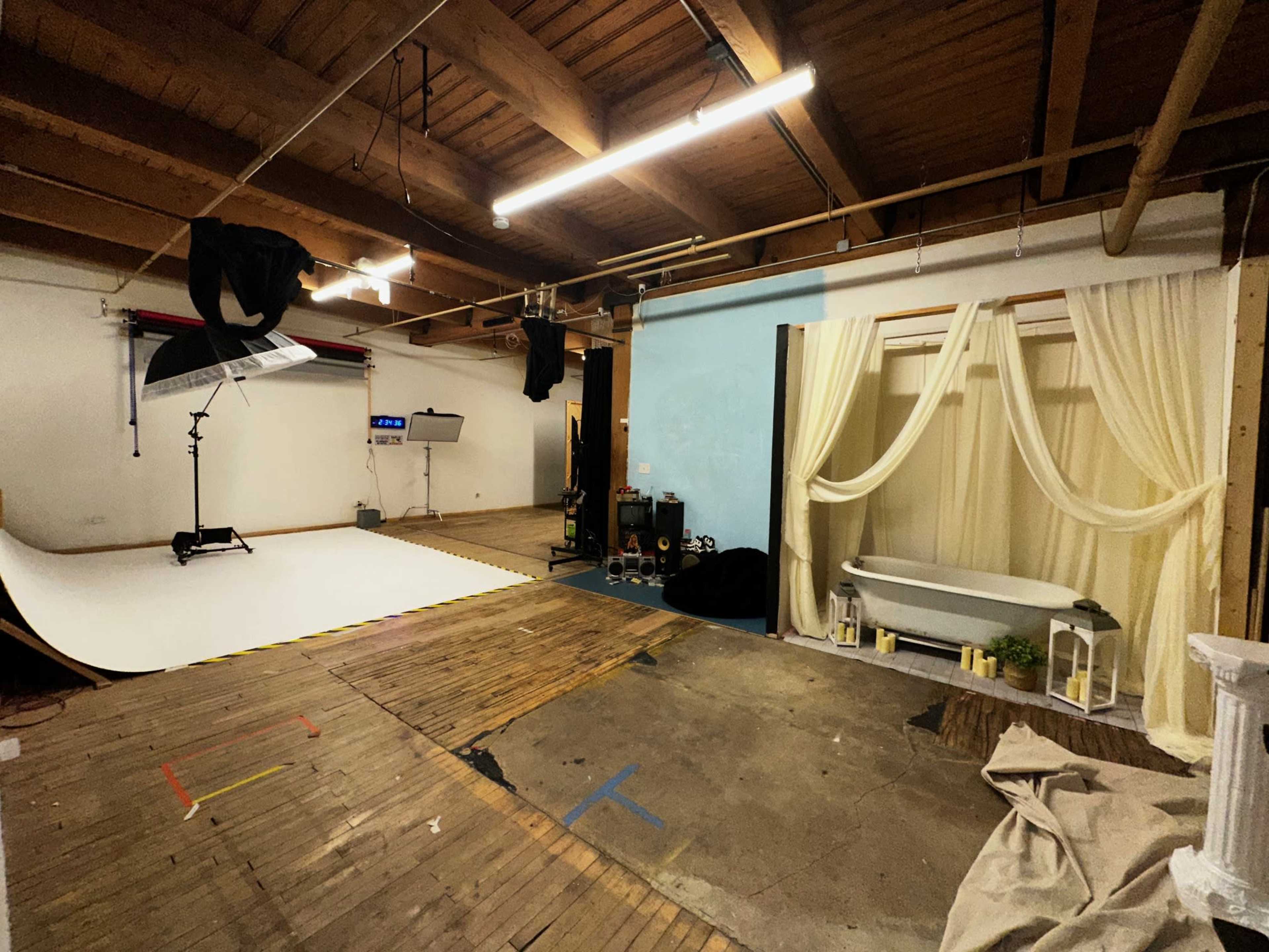 The image shows a spacious studio with wooden beams, a large white backdrop set up for photography, and a decorated area featuring a bathtub and draped curtains.