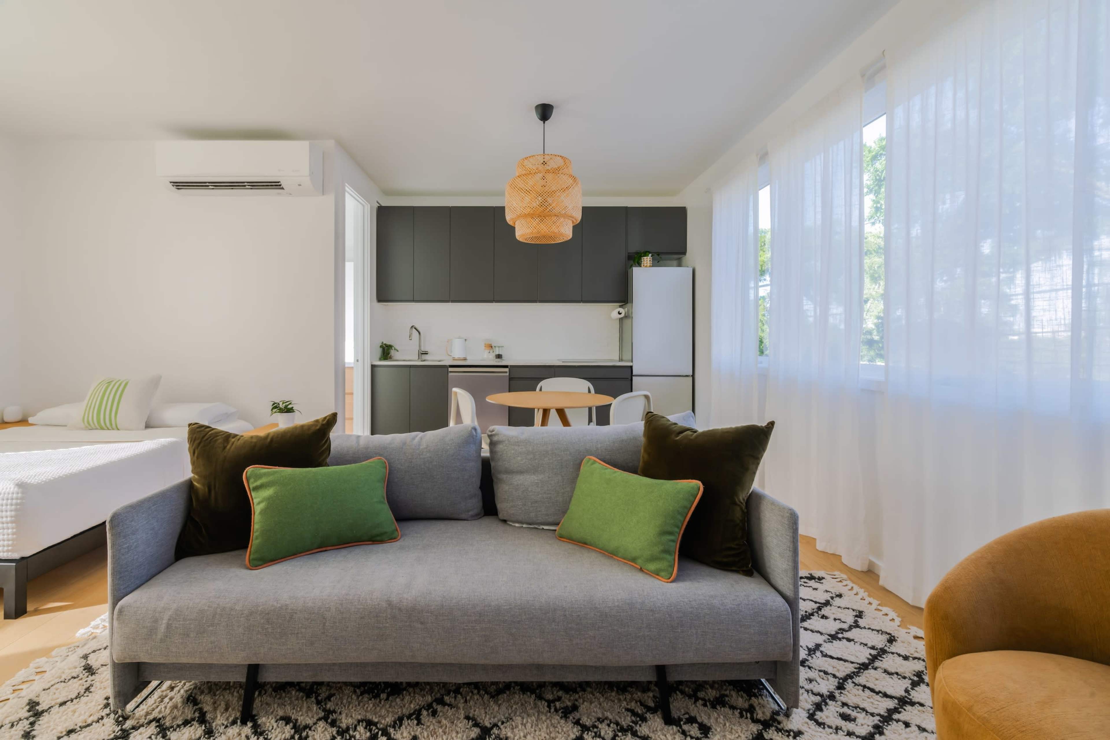 A modern living space features a gray sofa with green pillows, a small dining table, a kitchenette, and large windows allowing natural light.