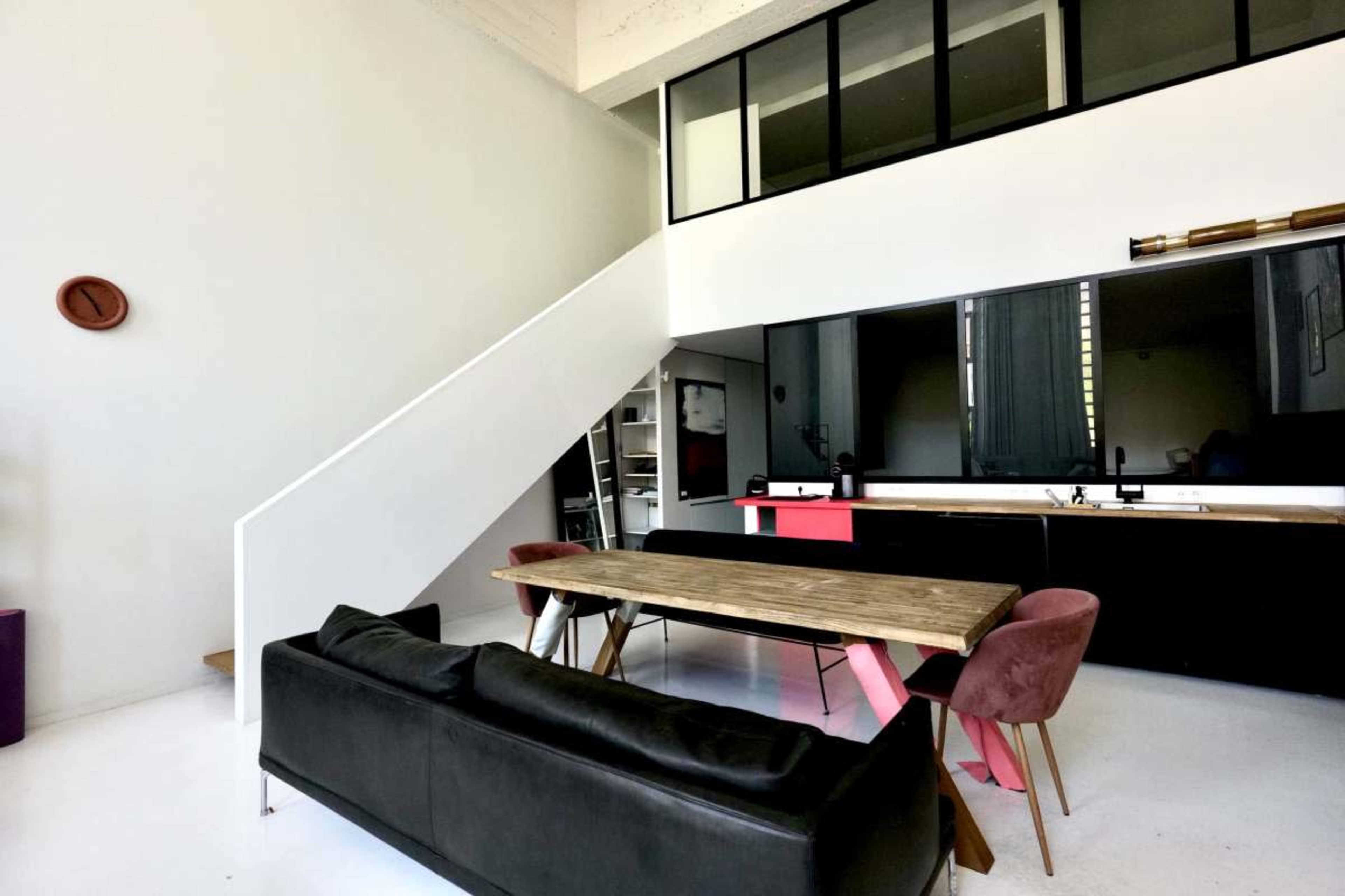 The image shows a modern, open-plan living space with a staircase, a wooden dining table surrounded by pink chairs, and a kitchen area featuring black cabinetry.