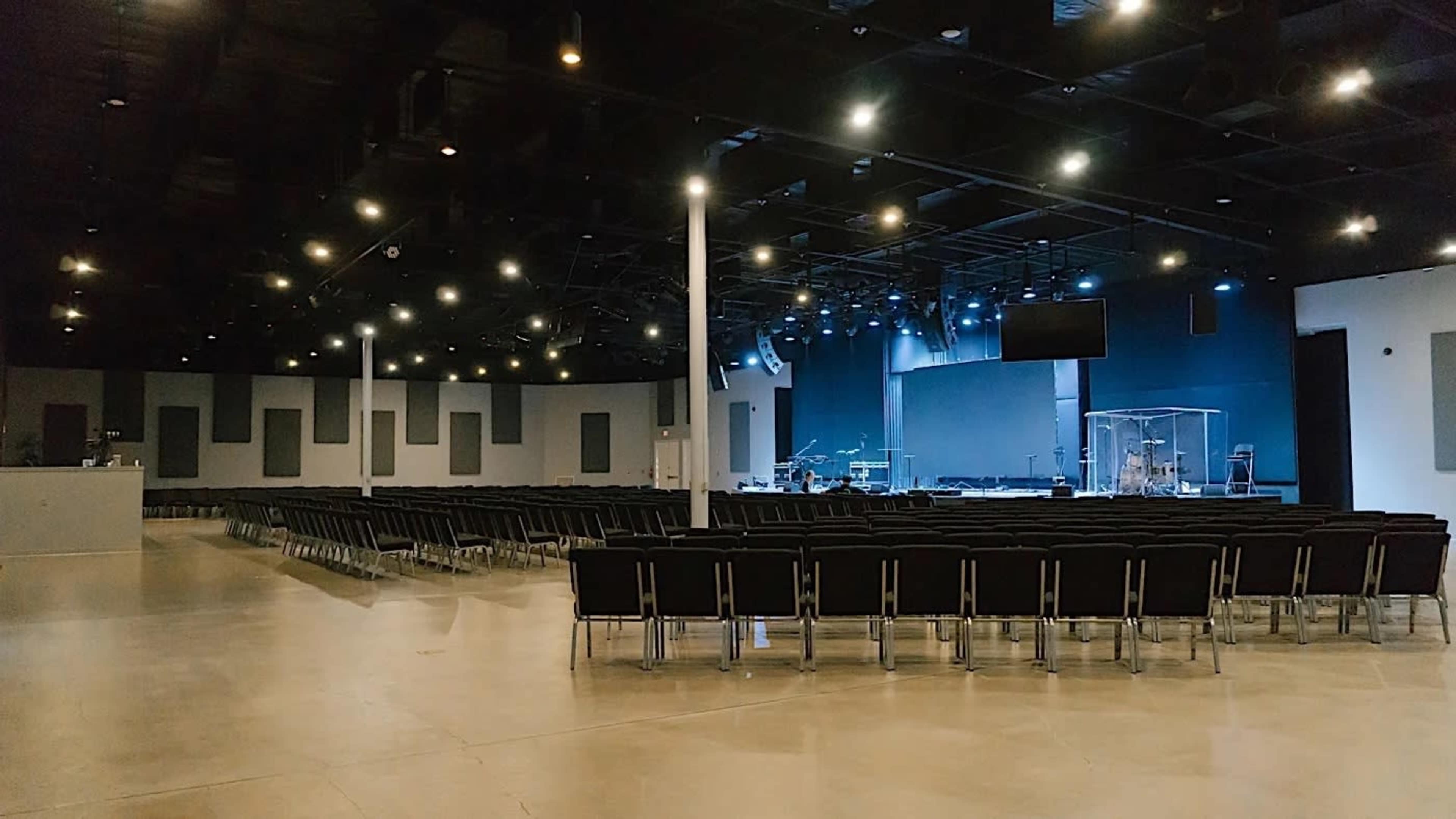 A spacious indoor auditorium with rows of black chairs facing a stage equipped with lighting and musical instruments.
