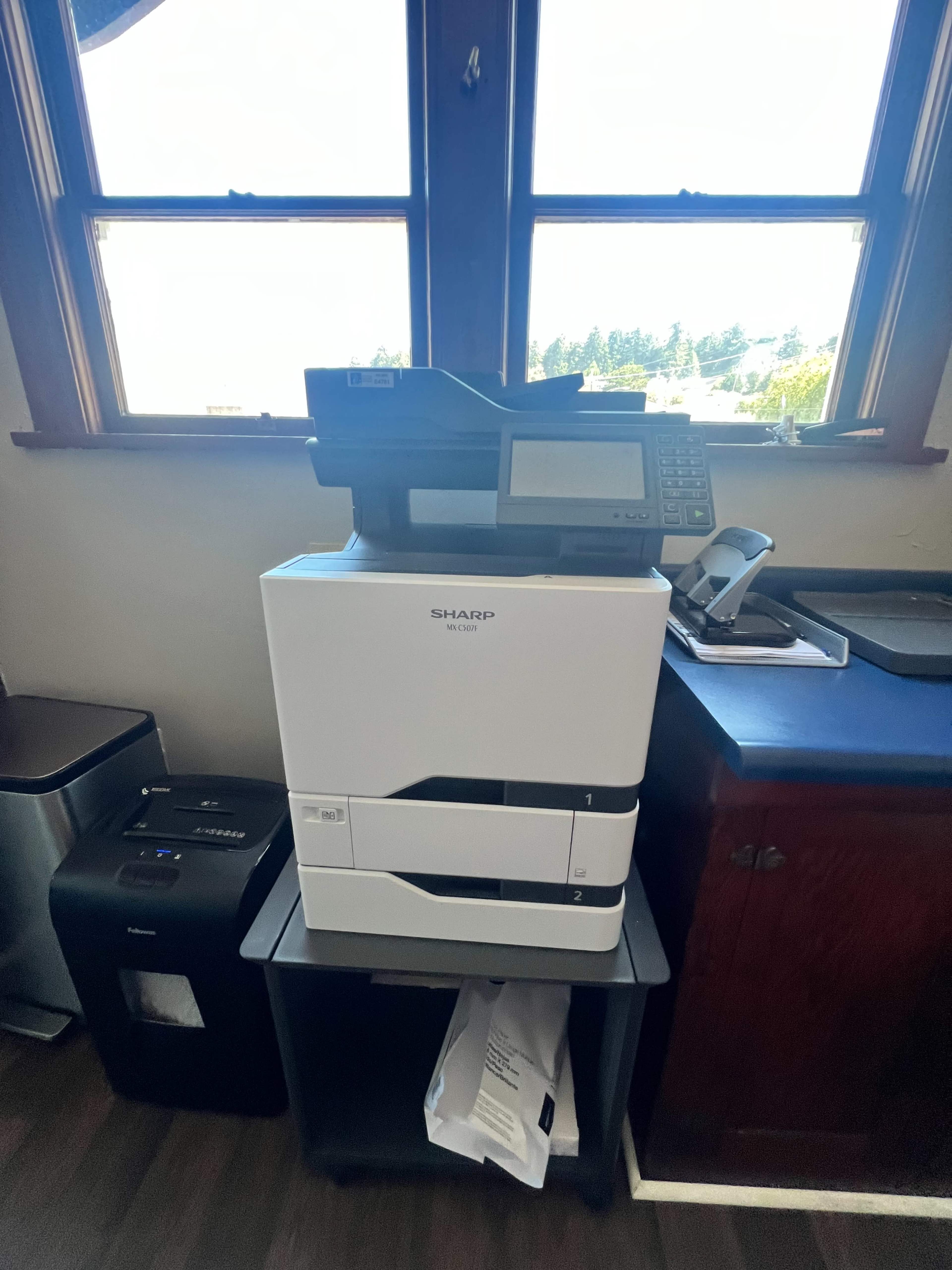 A multi-function printer is placed next to a desk with a small waste bin and a phone on top.