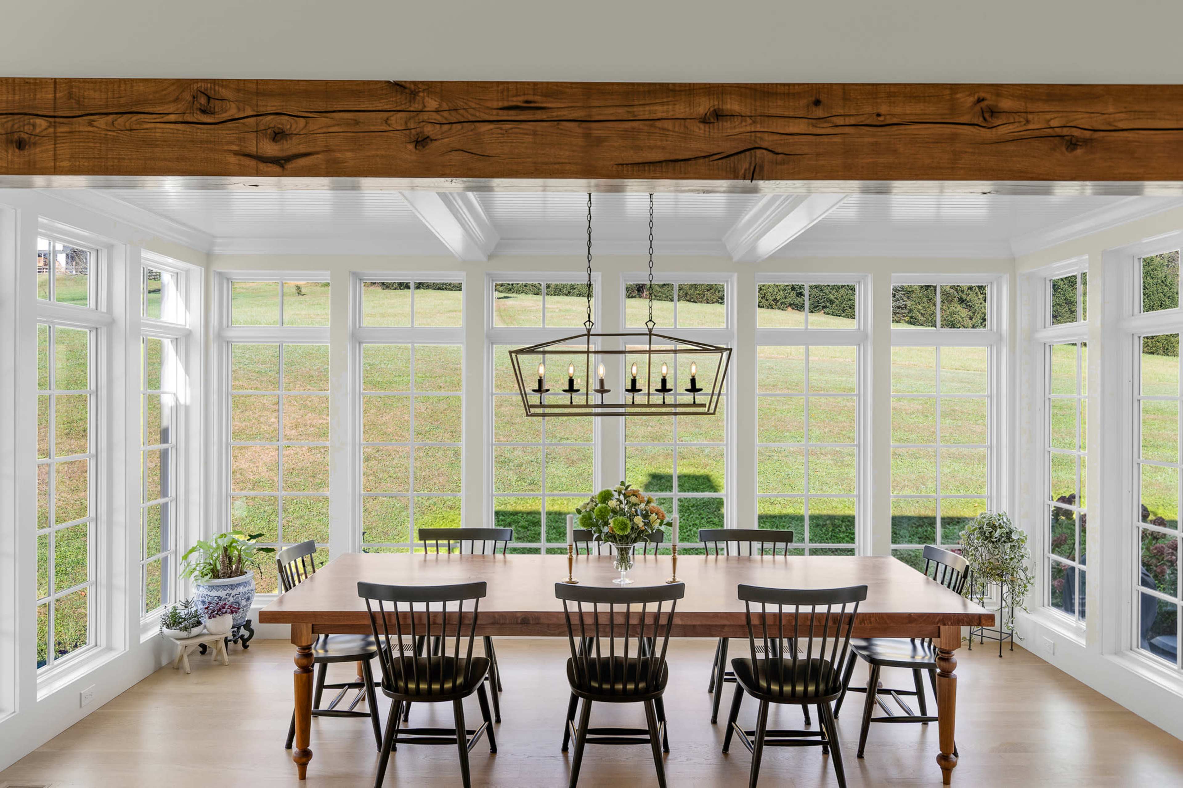 A spacious dining area features a large wooden table surrounded by black chairs, illuminated by a hanging light fixture, with multiple large windows offering a view of green fields outside.
