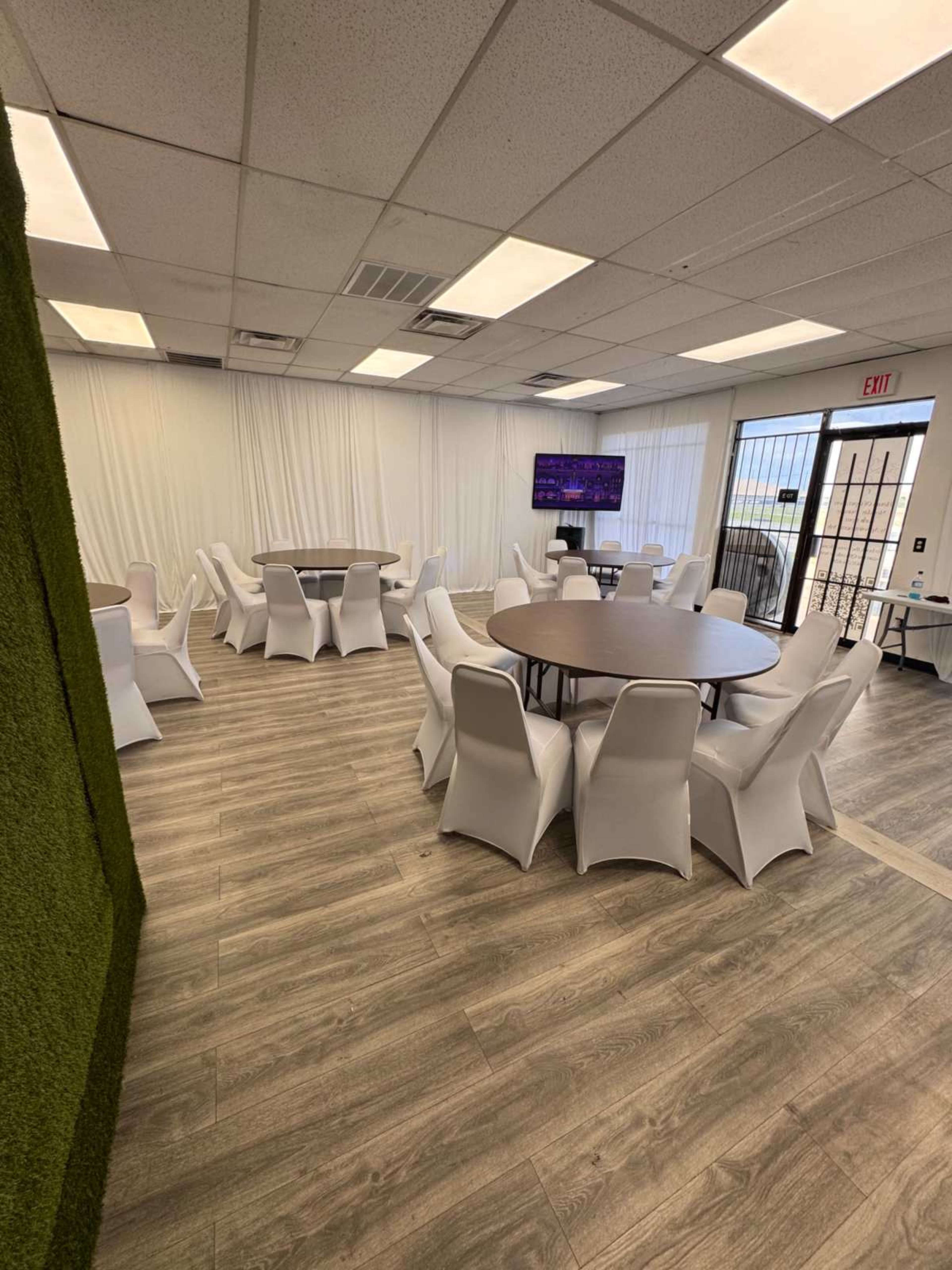 The image shows a brightly lit room with several round tables surrounded by white chairs, along with a green wall and a television displayed on one side.