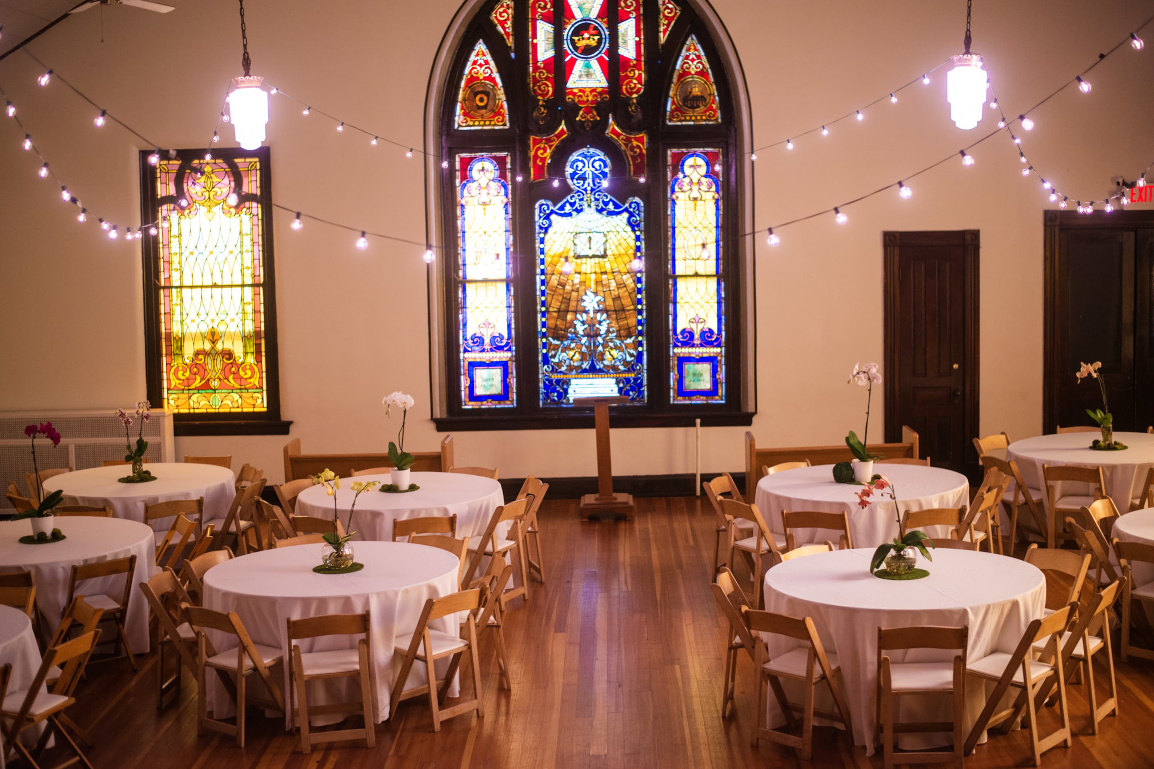 A spacious hall with round tables covered in white tablecloths is illuminated by string lights and features a large stained glass window at the back.