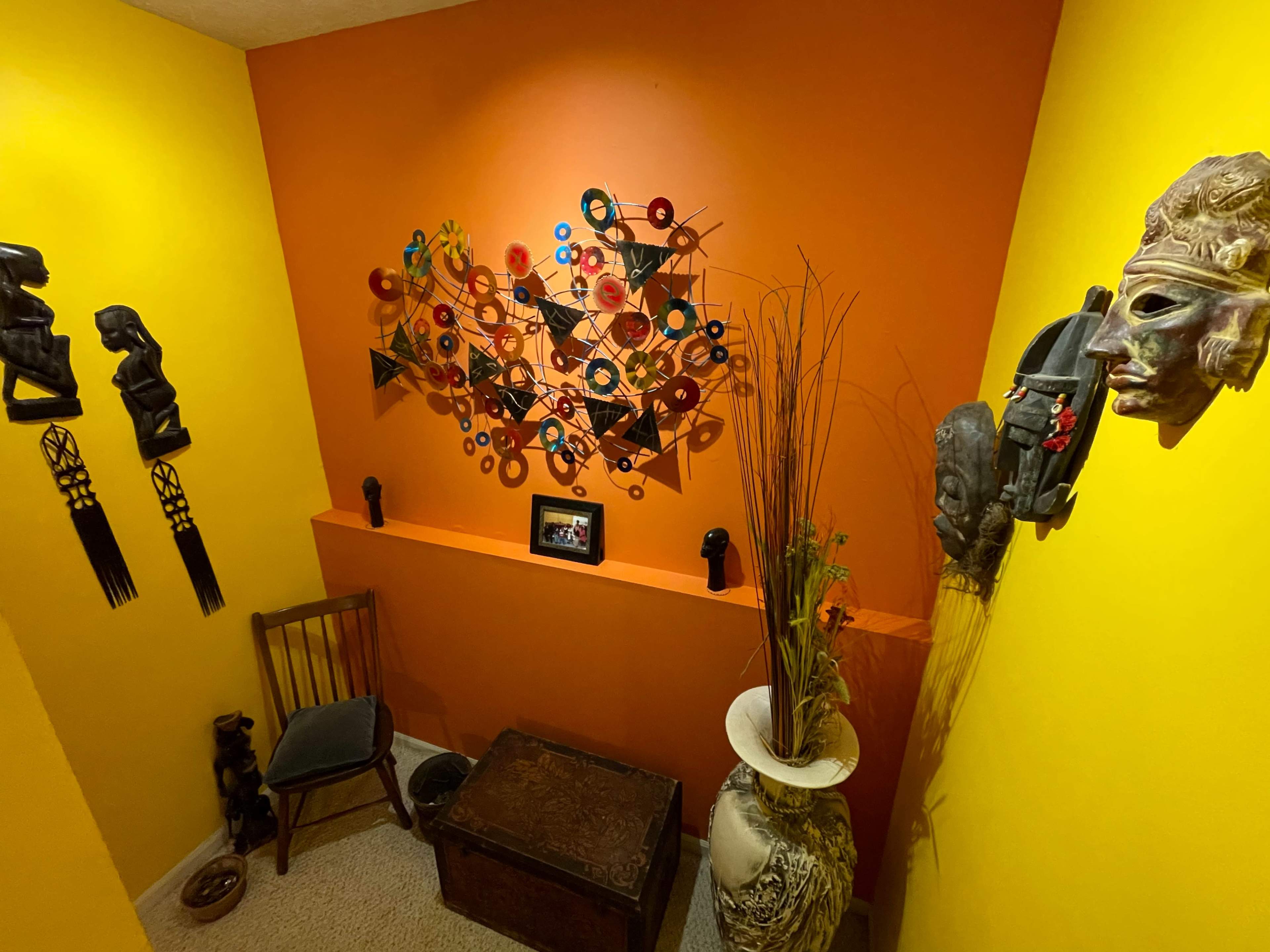The image shows a corner of a room with vibrant orange and yellow walls, featuring decorative wall art, masks, a wooden chair, a potted plant, and an ornate trunk.