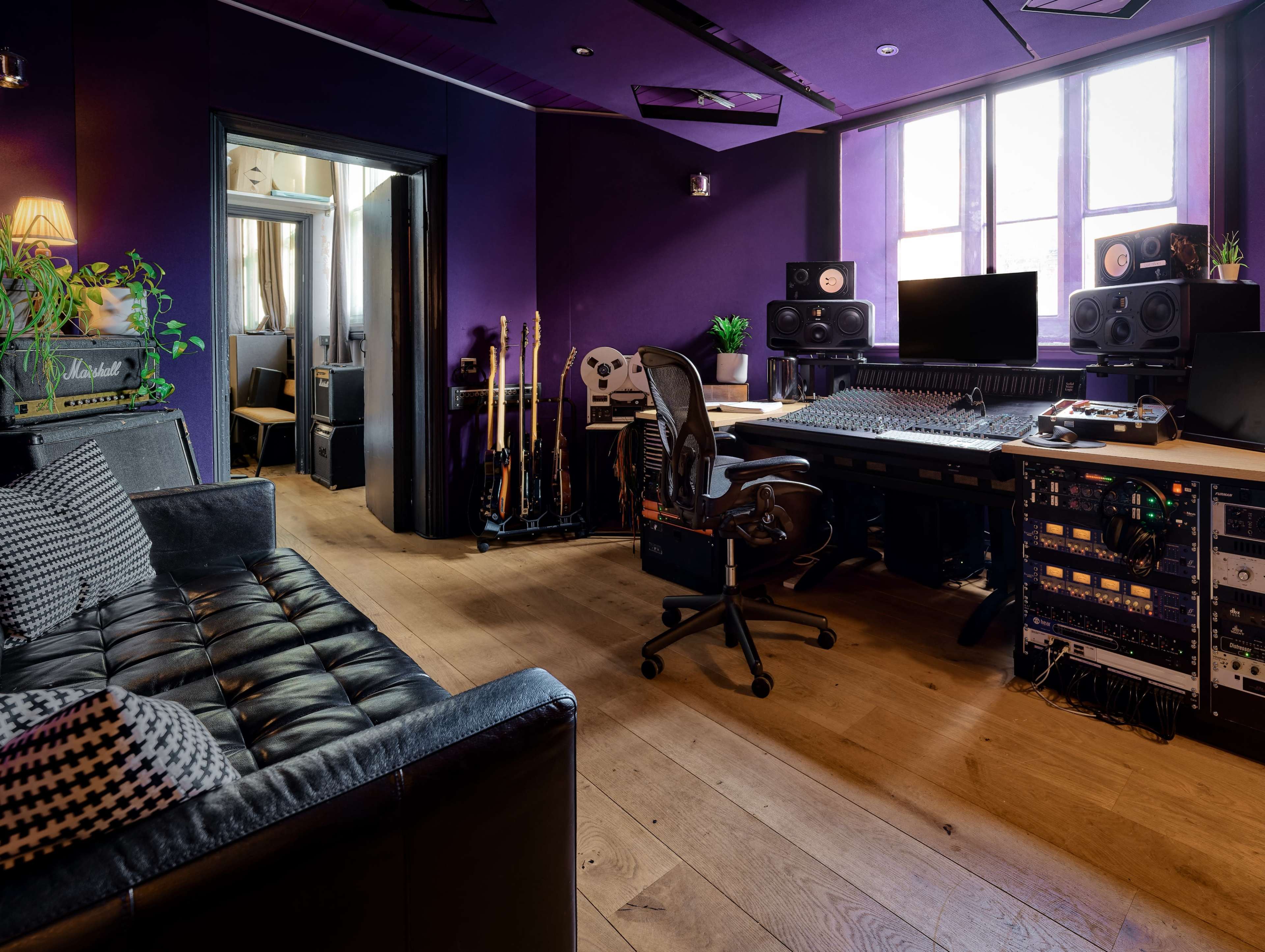 The image shows a music studio with purple walls, a large mixing desk, audio speakers, and a comfortable couch.