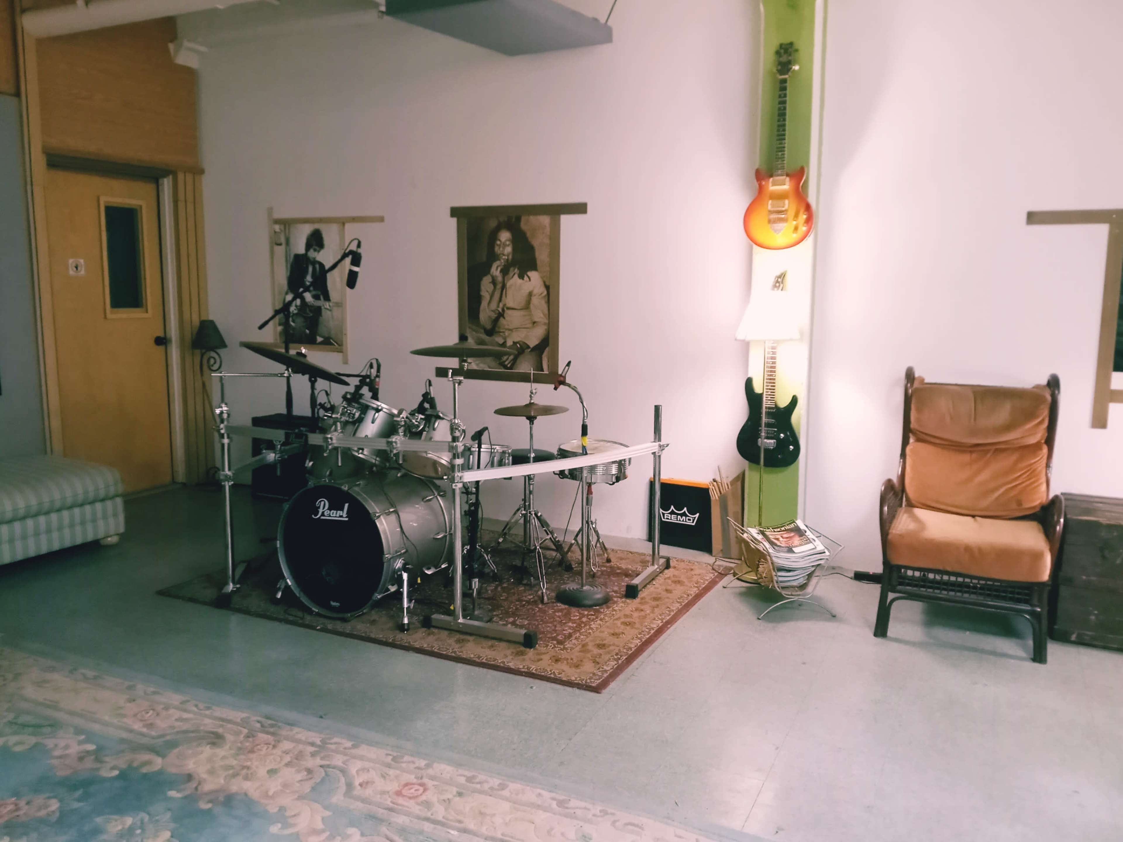 A music practice room featuring a drum set, electric guitars mounted on the wall, and a chair beside a lamp.