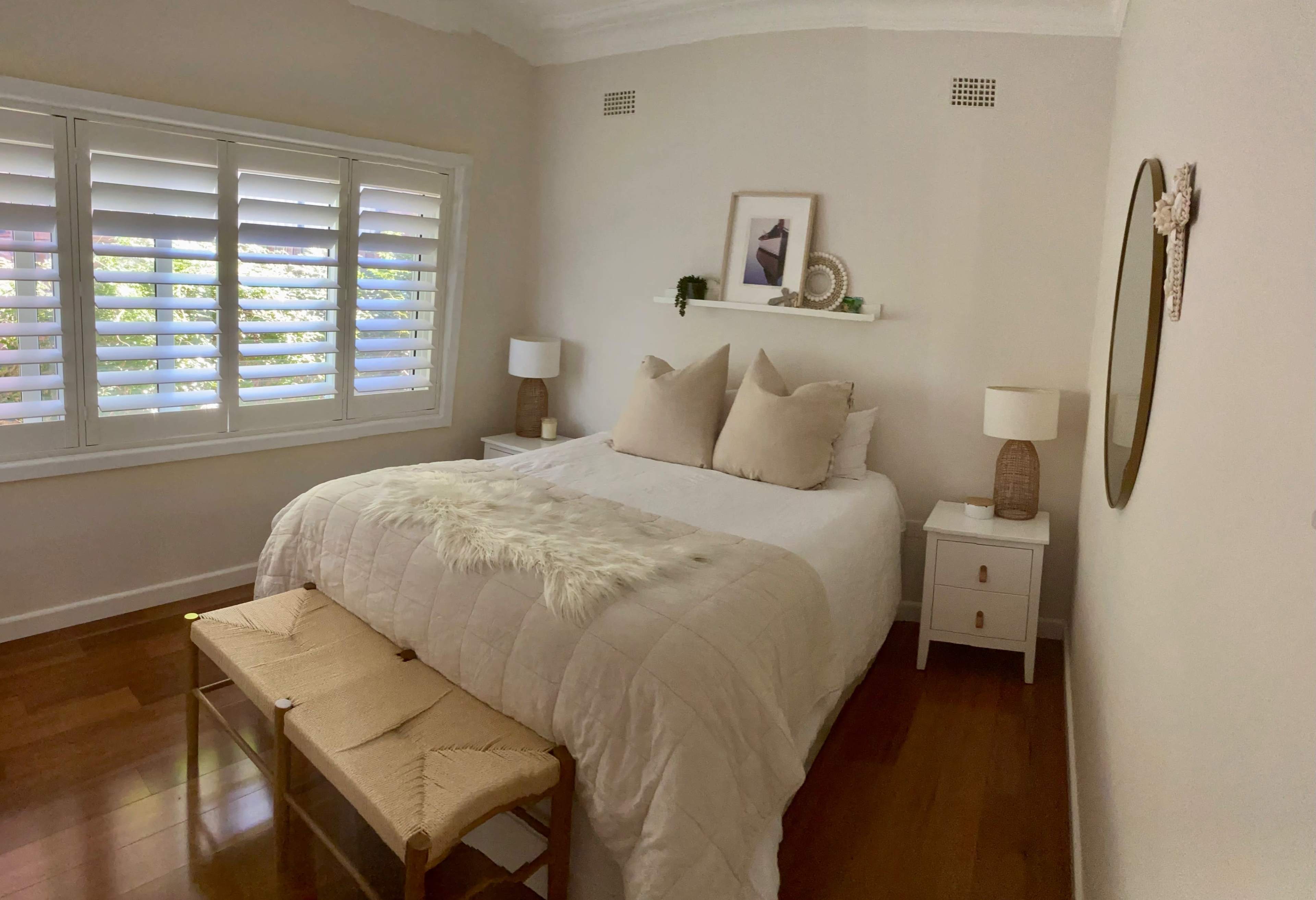 A neatly arranged bedroom features a bed with light-colored linens, two bedside tables with lamps, a bench at the foot of the bed, and decorative items on a shelf.