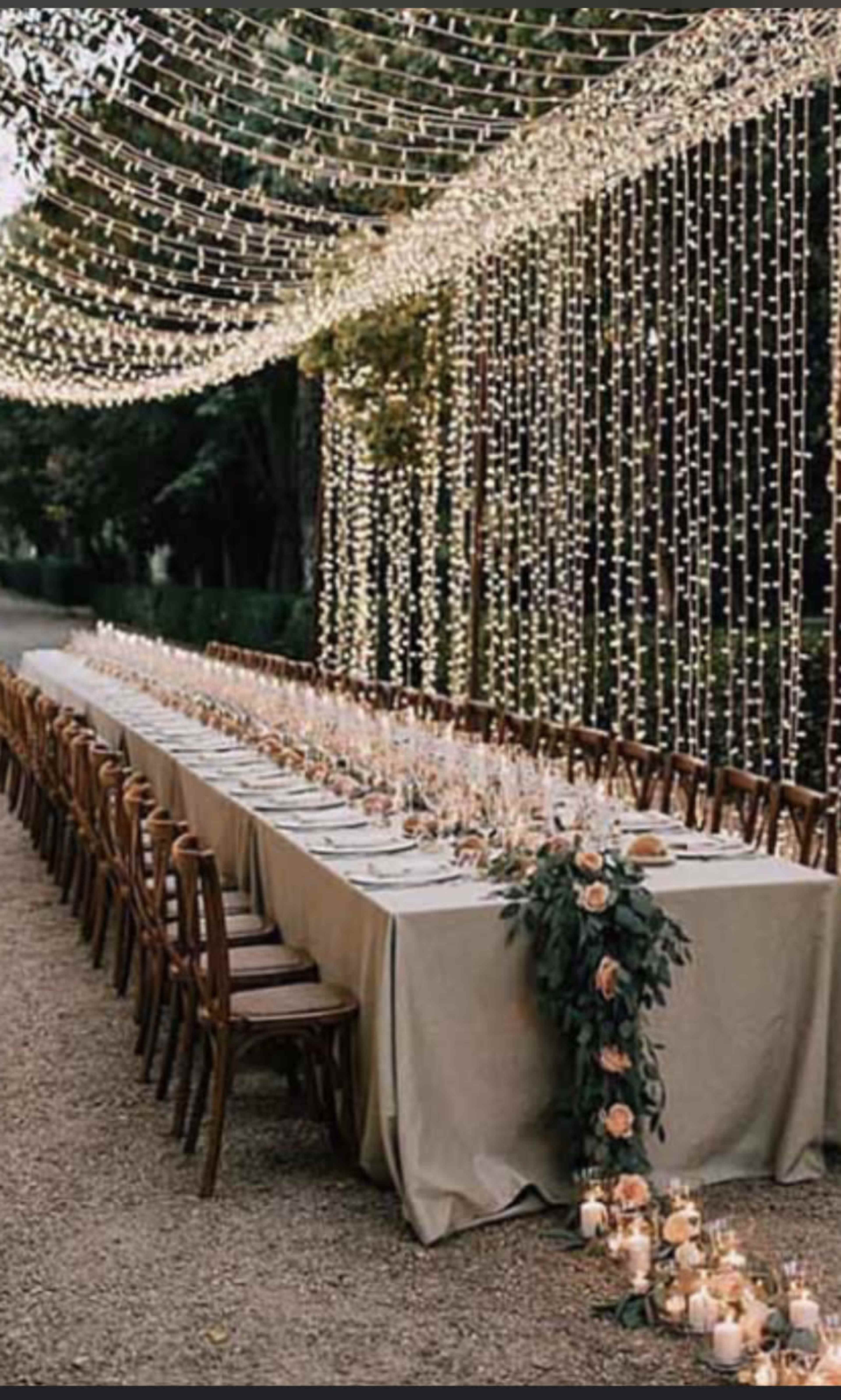 A long outdoor dining table is set up under a canopy of string lights, surrounded by wooden chairs and adorned with floral arrangements and candles.