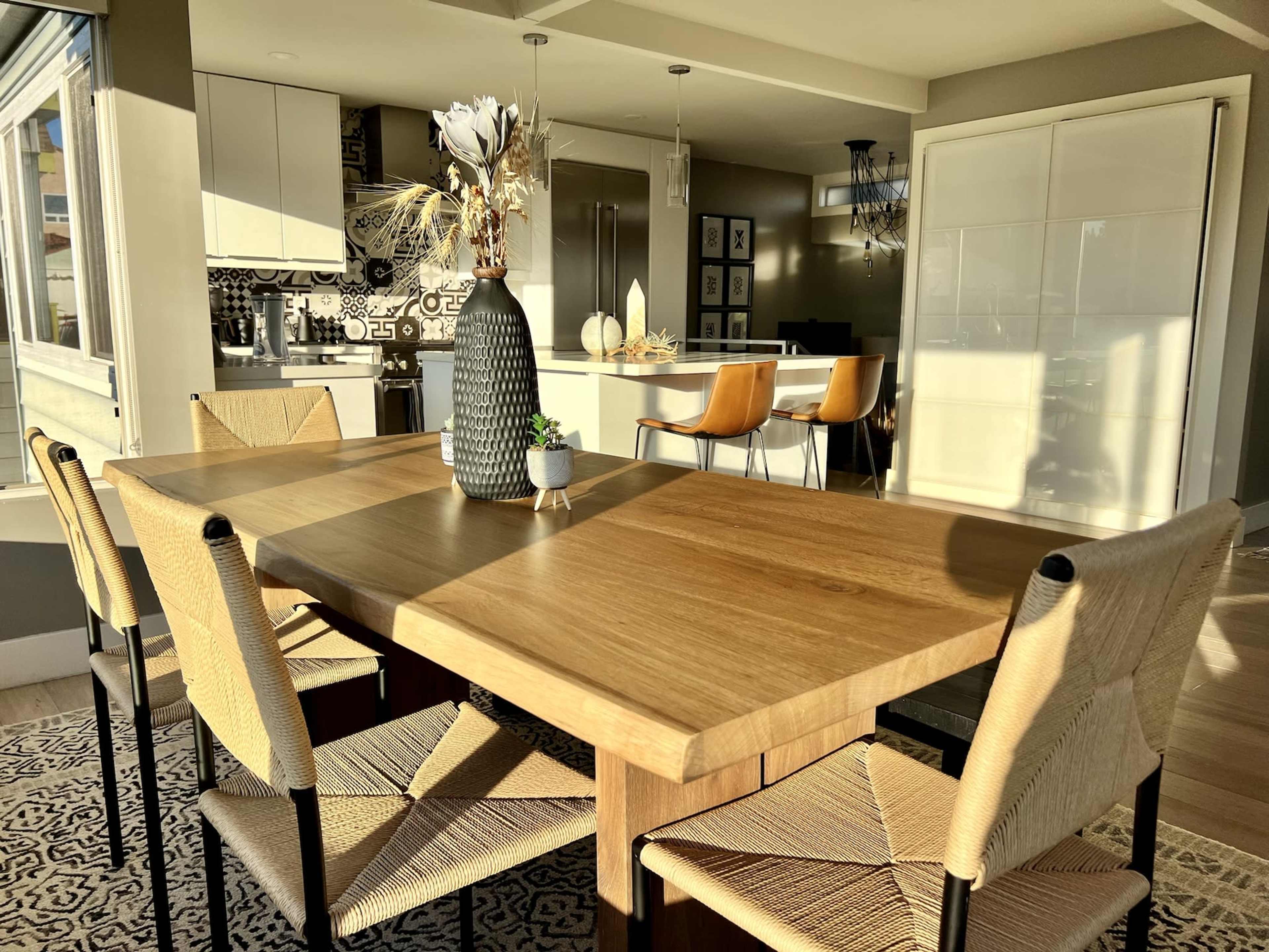 A wooden dining table with woven chairs is set in a brightly lit room featuring a modern kitchen in the background.