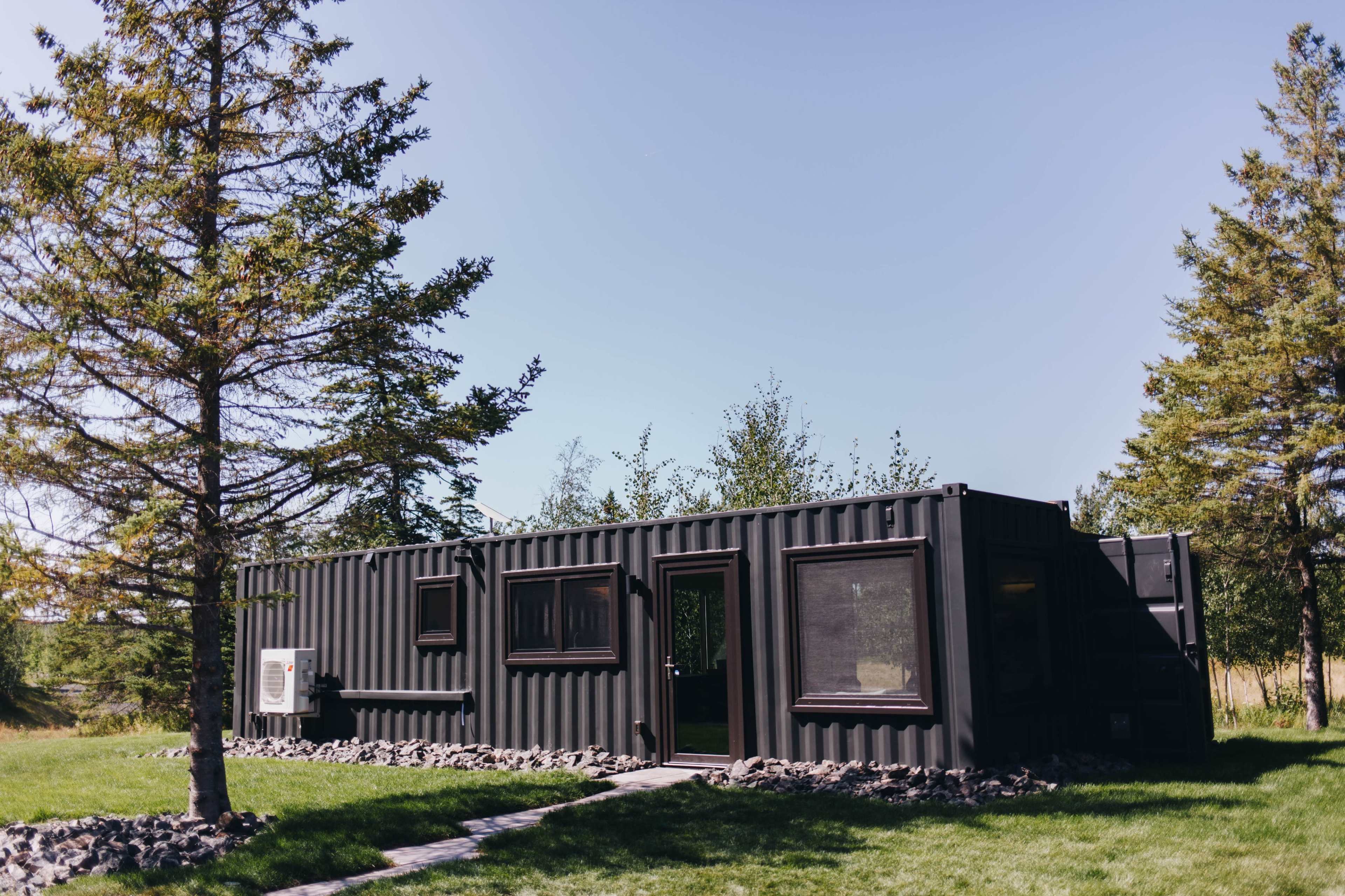 A black container home is situated amid trees and grass, with a clear blue sky overhead.