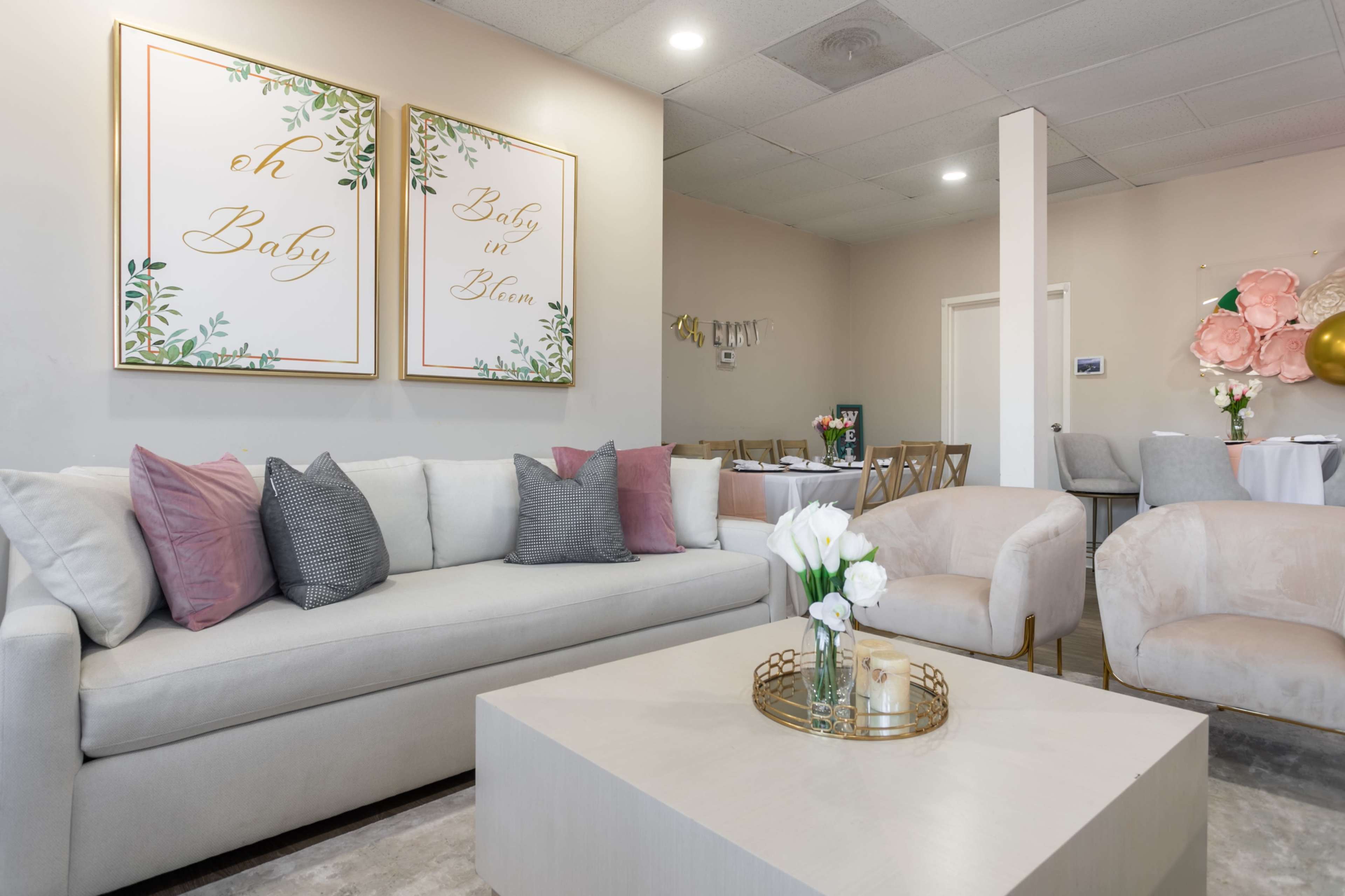A well-decorated lounge area featuring a light-colored sofa with decorative pillows, a coffee table with a tray, and framed wall art with text related to baby celebrations.
