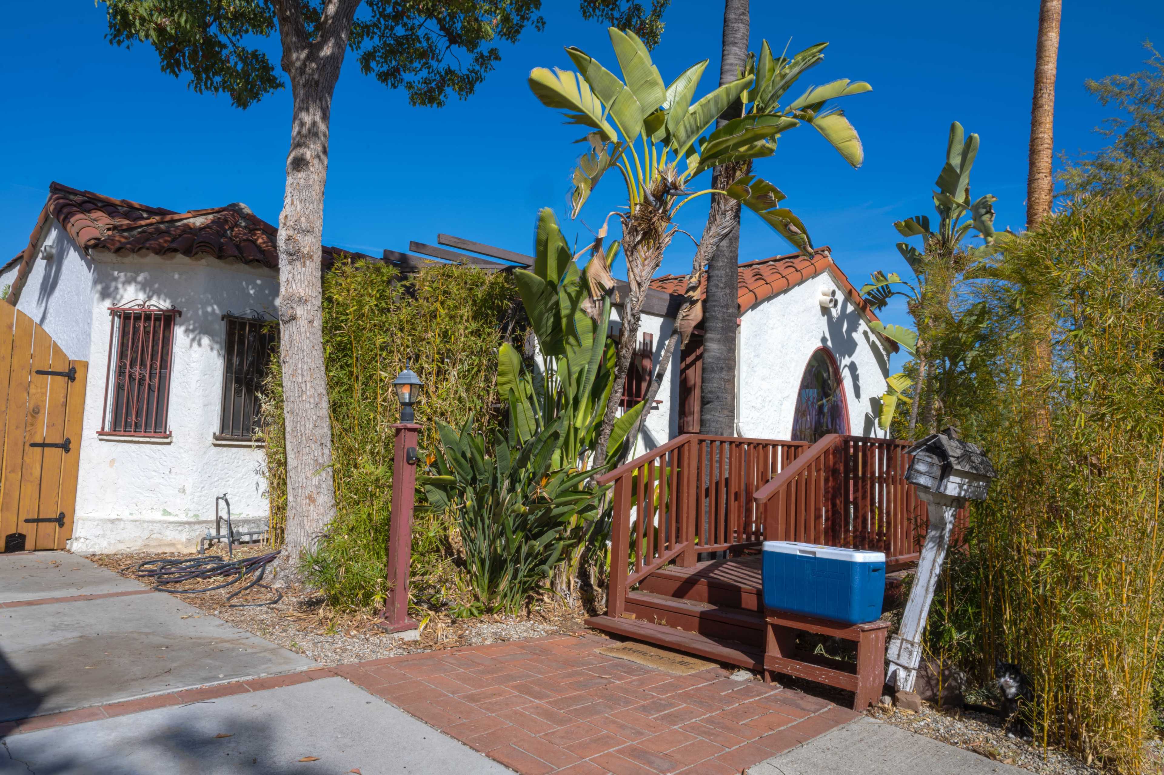 A wooden ramp leads to a doorway of a white stucco house surrounded by palm trees and greenery.
