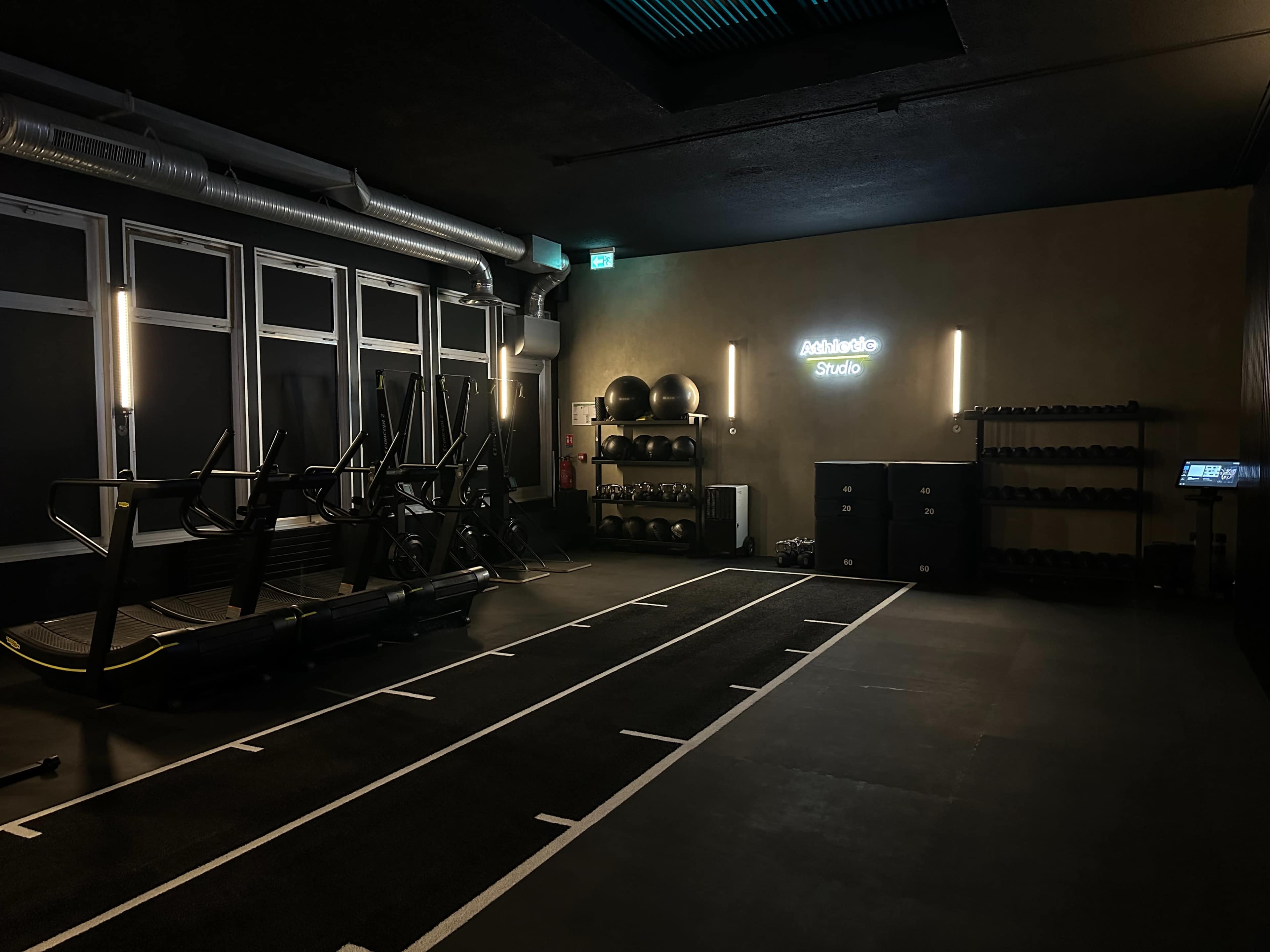 The image shows a dimly lit gym studio equipped with exercise machines, weights, and fitness accessories, with a neon sign reading "Athletic Studio" on the wall.
