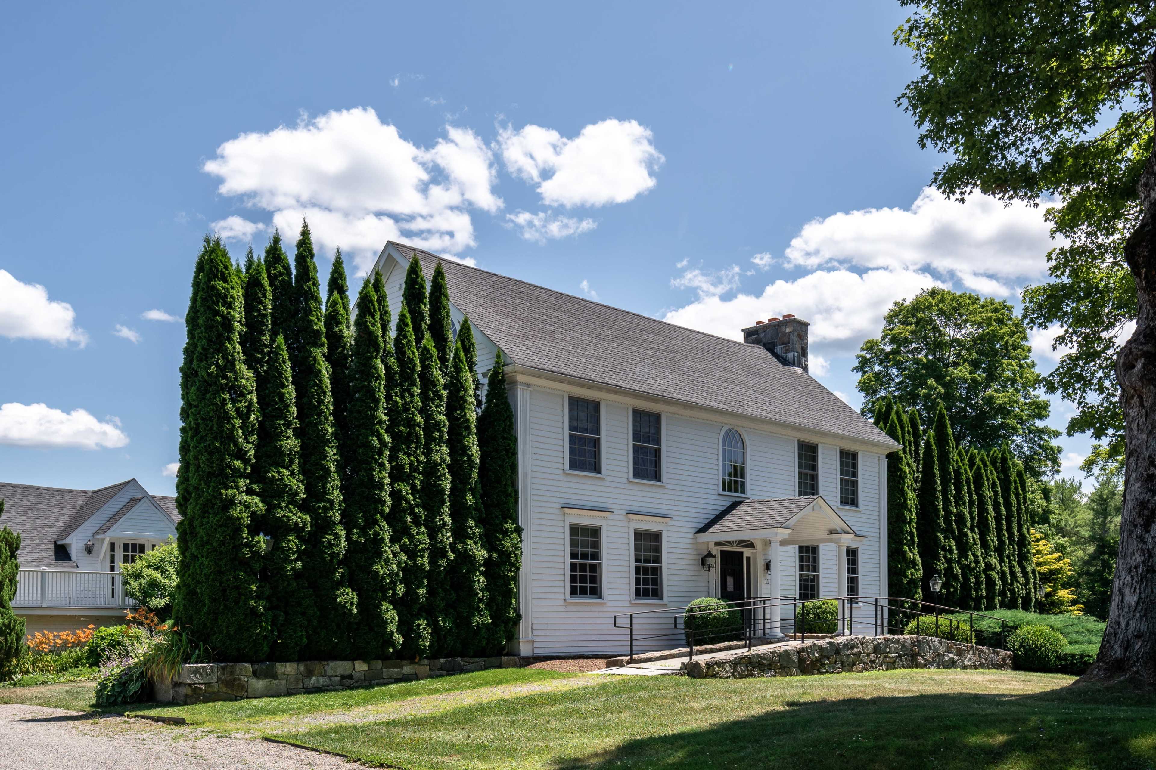 A two-story white house with a steep roof is surrounded by tall evergreen trees and a neatly manicured lawn.