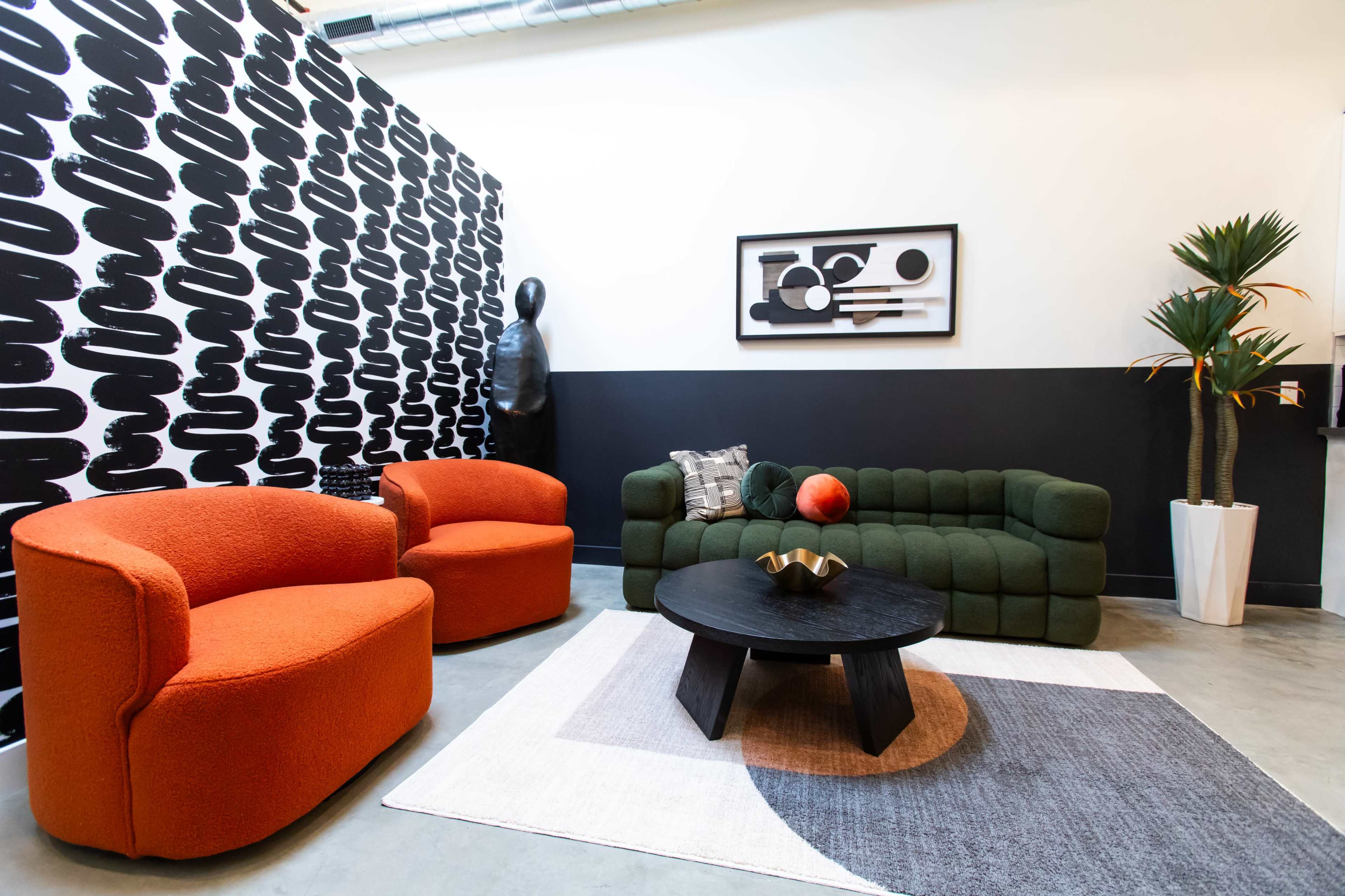 A modern seating area features two orange armchairs and a green sofa arranged around a round black coffee table on a patterned rug, against a black and white patterned wall.