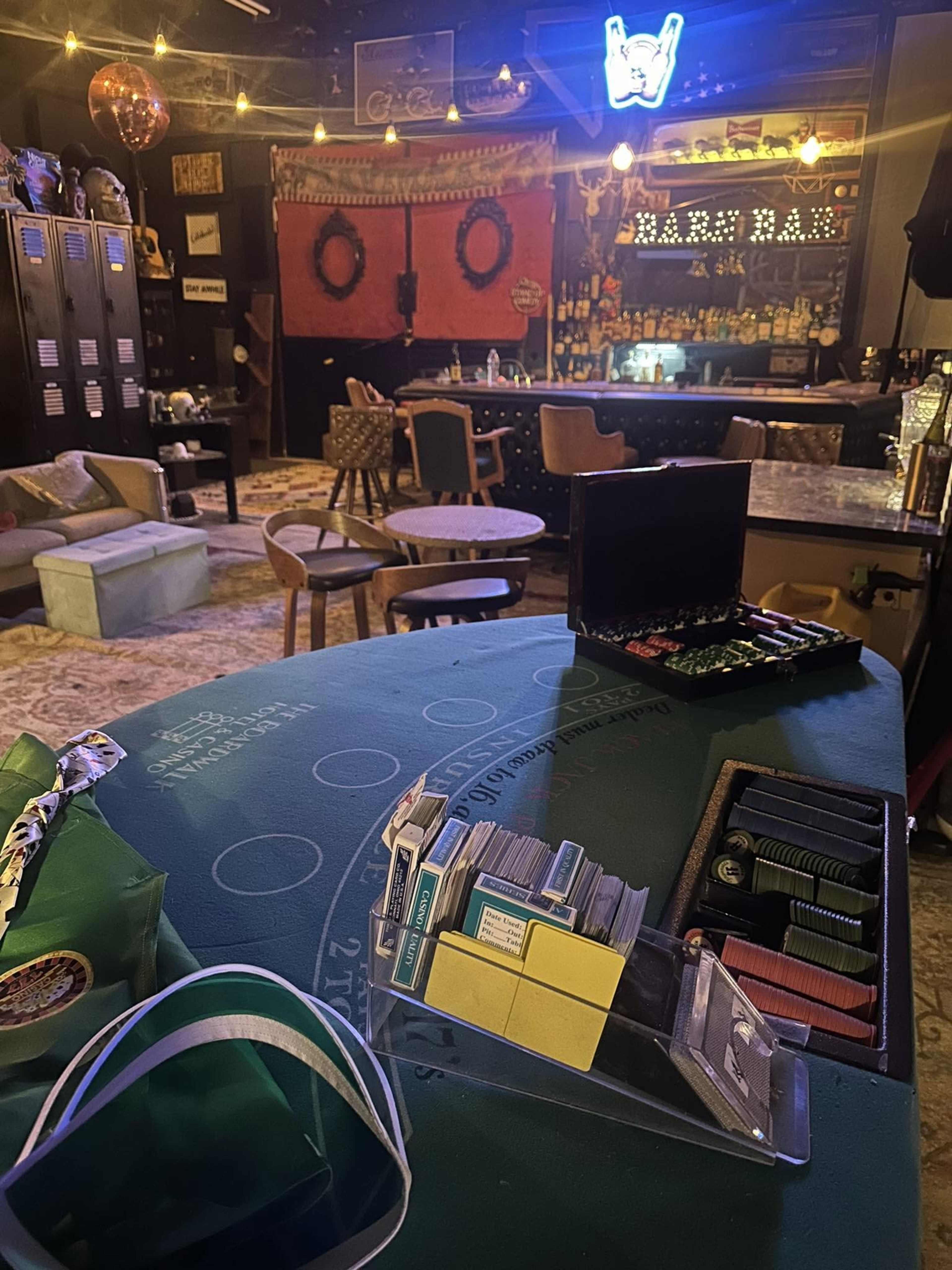 The image shows a low-lit gaming area with a green felt table featuring poker chips and cards, surrounded by various tables and chairs in a casual bar setting.
