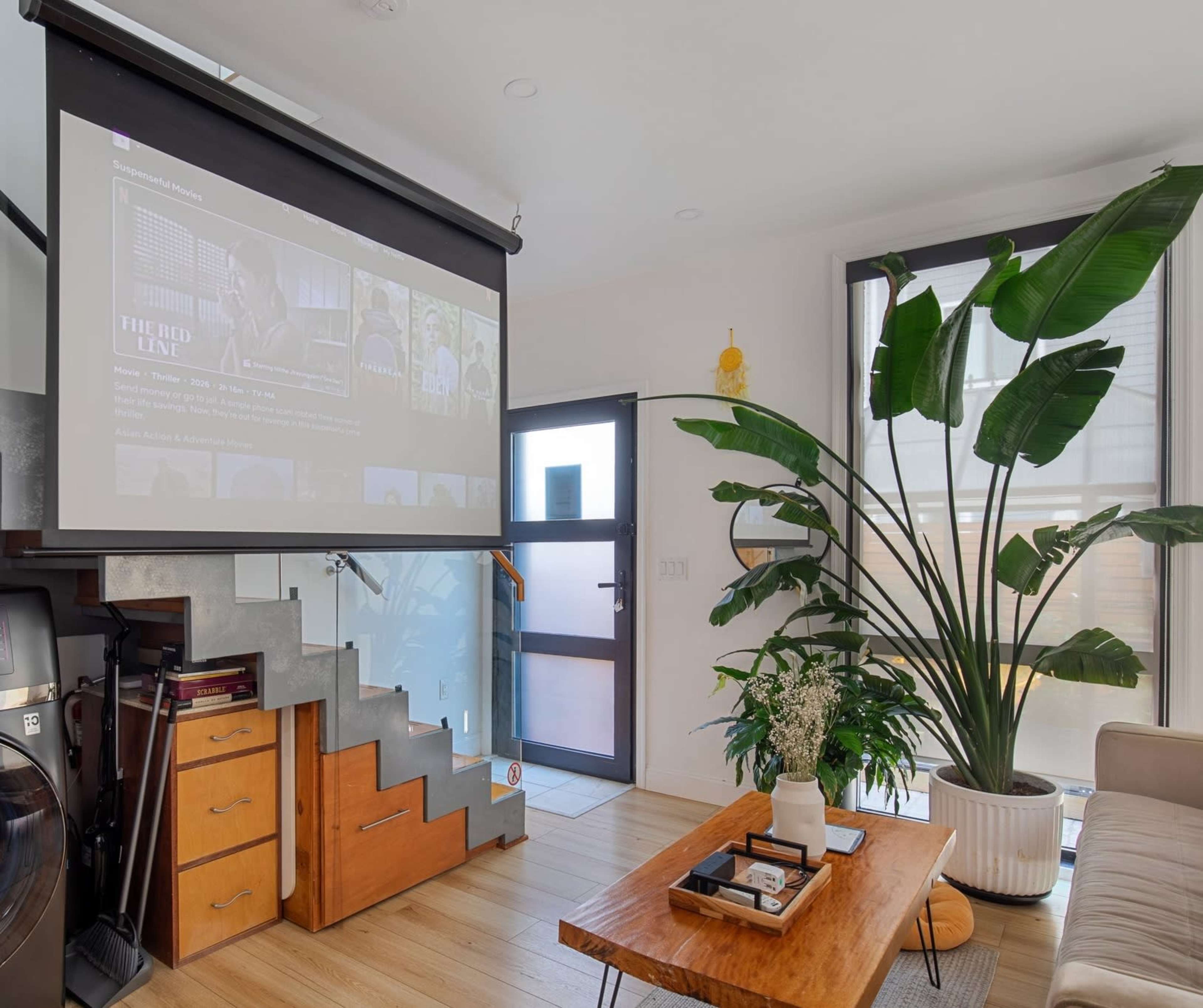 The image shows a modern living space featuring a large projecting screen displaying content, a wooden coffee table, a tall indoor plant, and a staircase in the background.