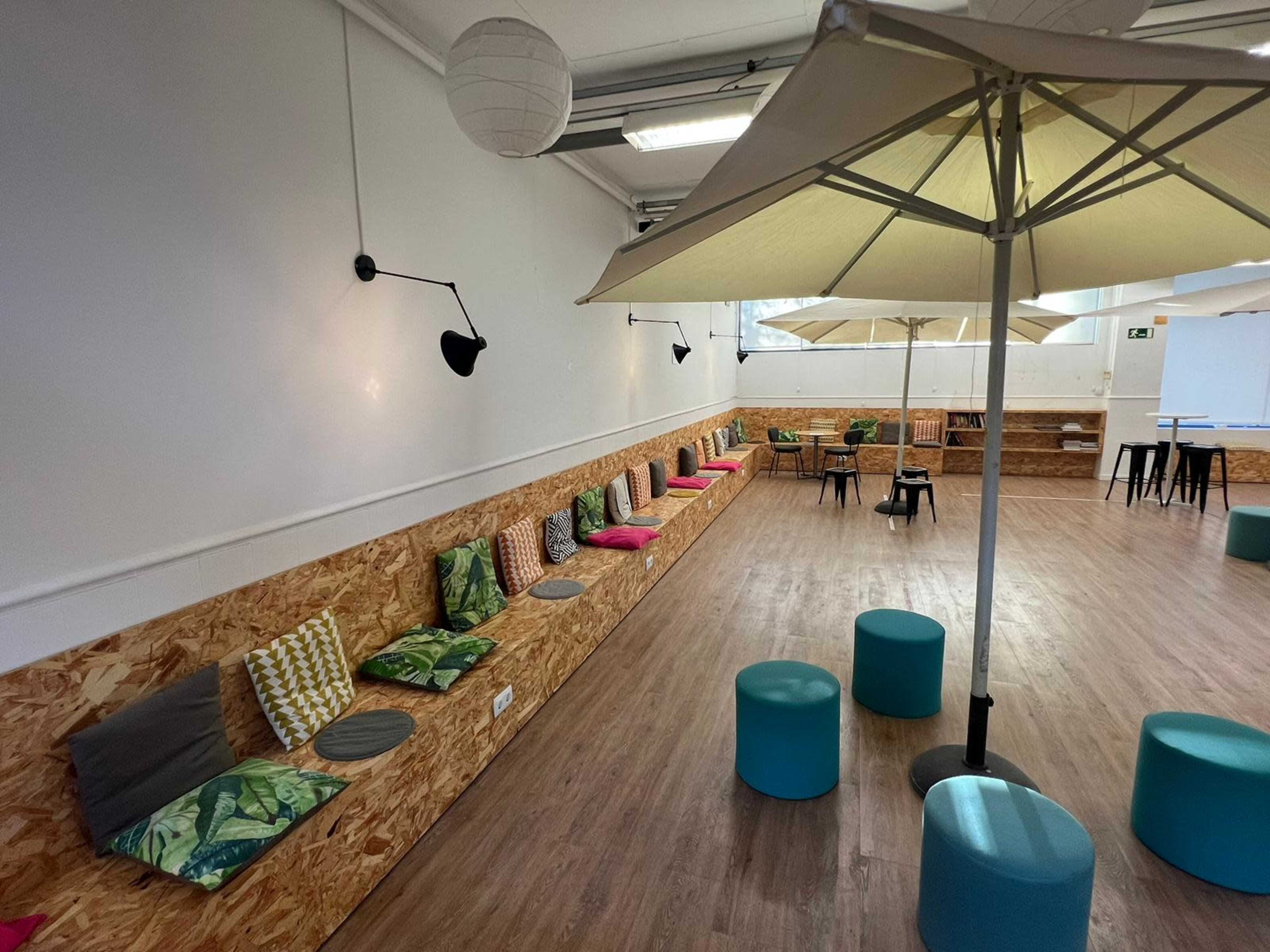 A spacious room with a long wooden bench featuring colorful cushions and several round stools, illuminated by wall-mounted lamps and large umbrellas.