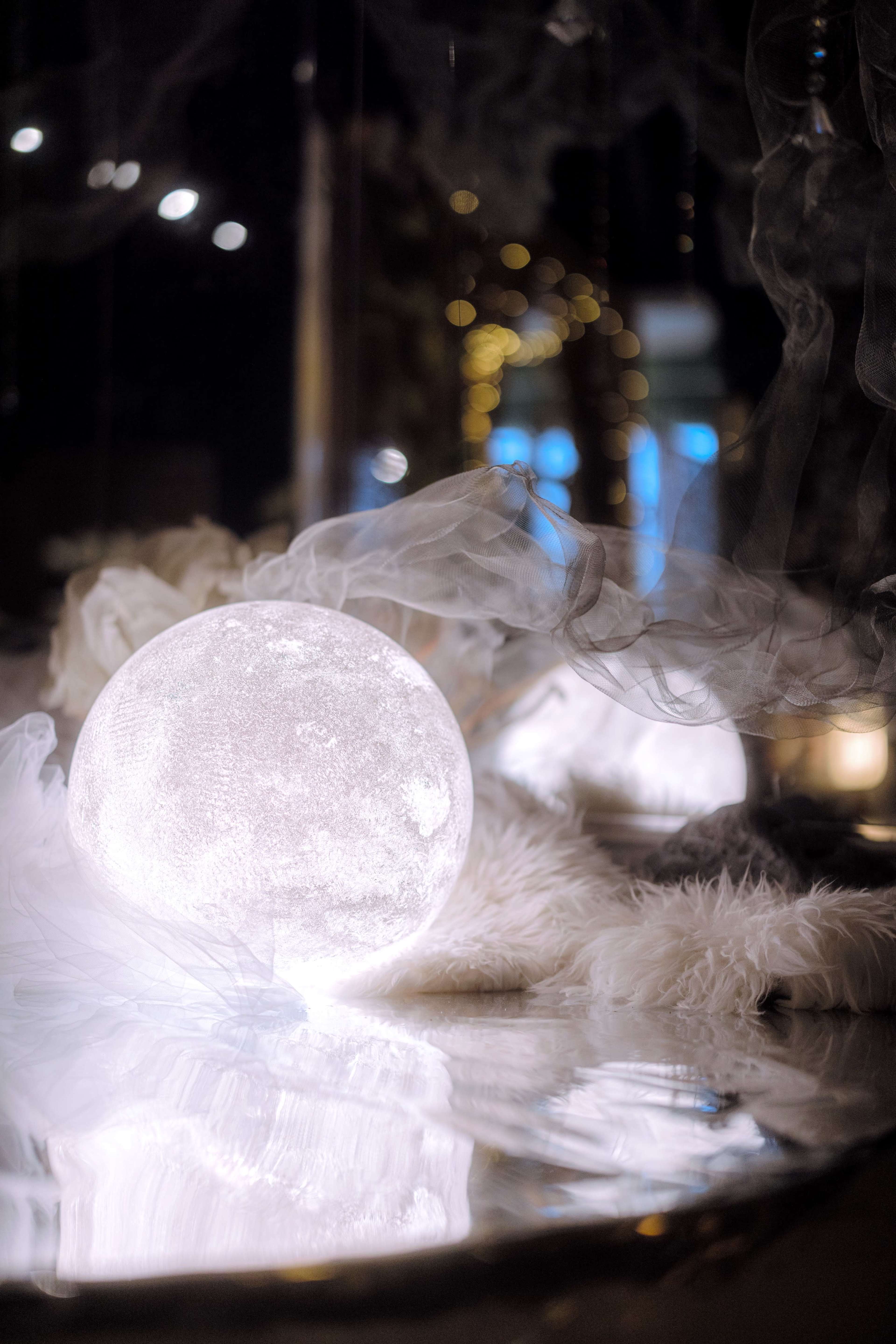 A glowing sphere resembling the moon casts light on a reflective surface surrounded by soft textures and wisps of fog.