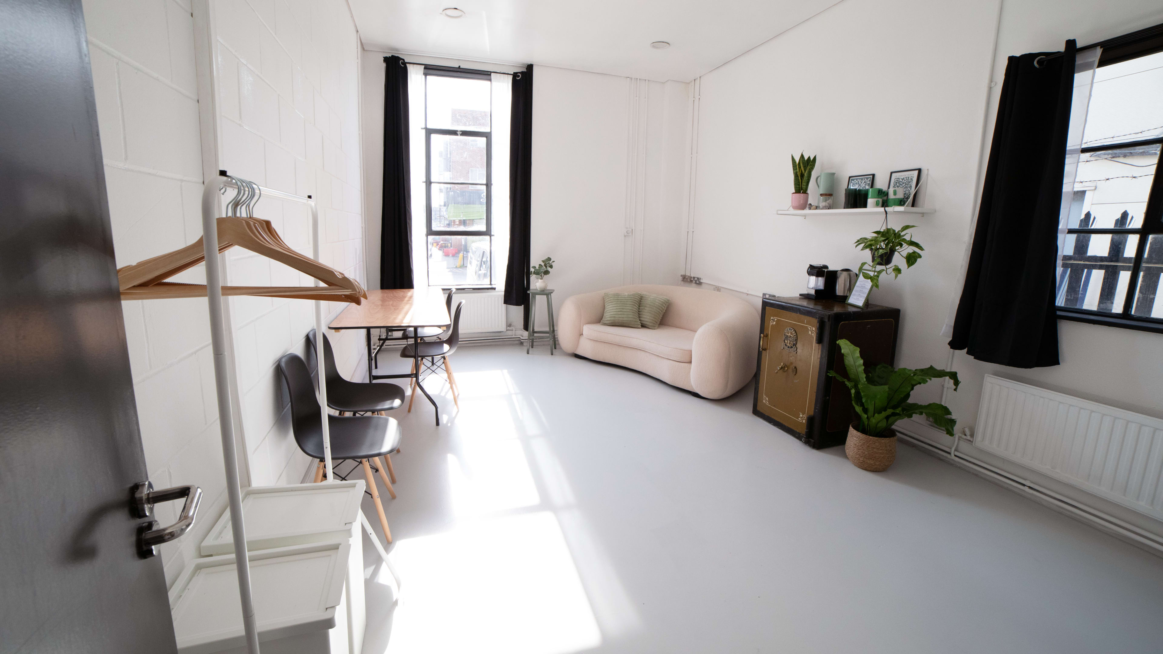 The room features a sofa, a table with chairs, a small cabinet, and several plants, all arranged under large windows with natural light.