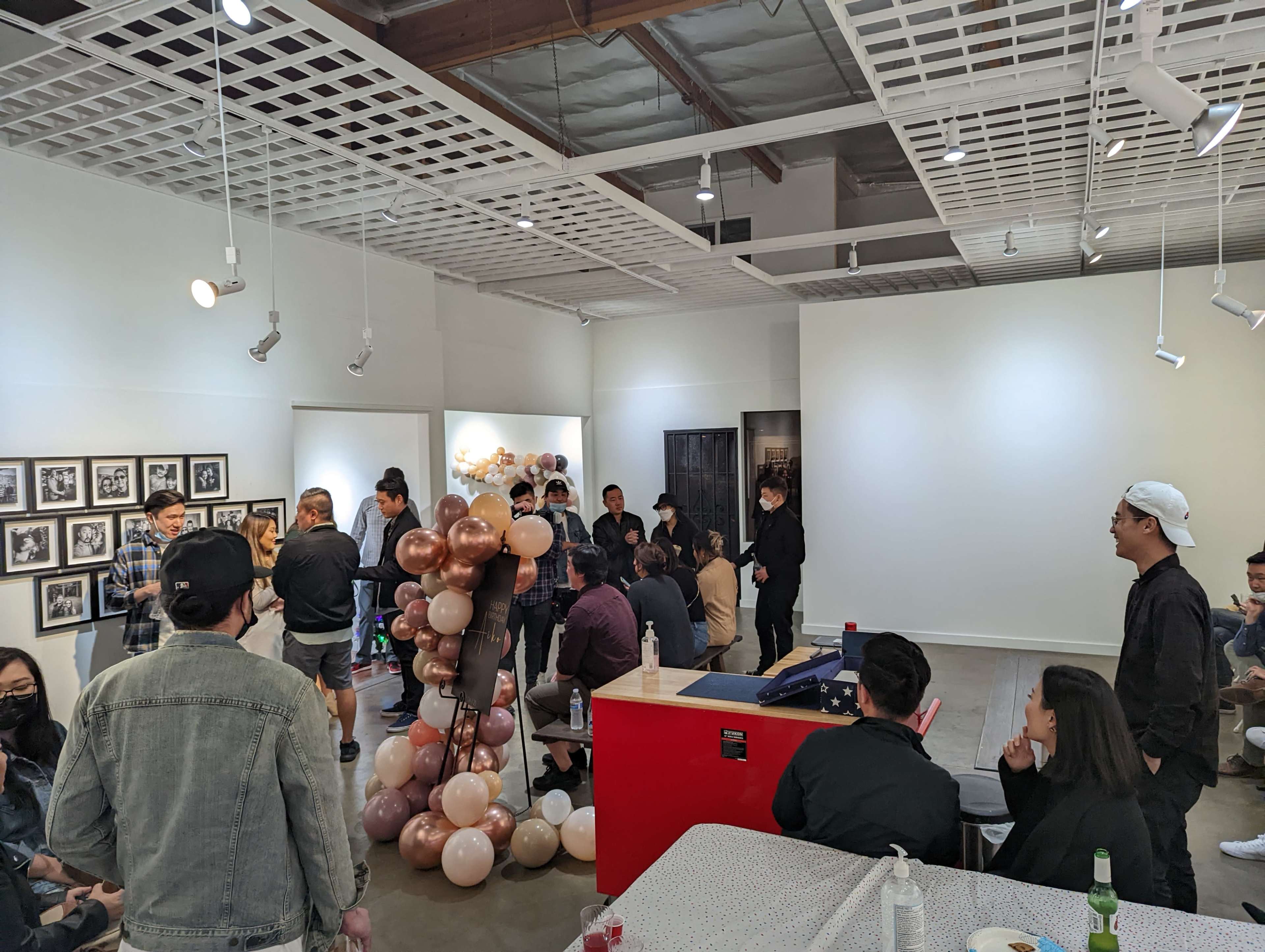 A group of people are gathered in an art gallery, mingling around a decorative balloon column and various artworks displayed on the walls.