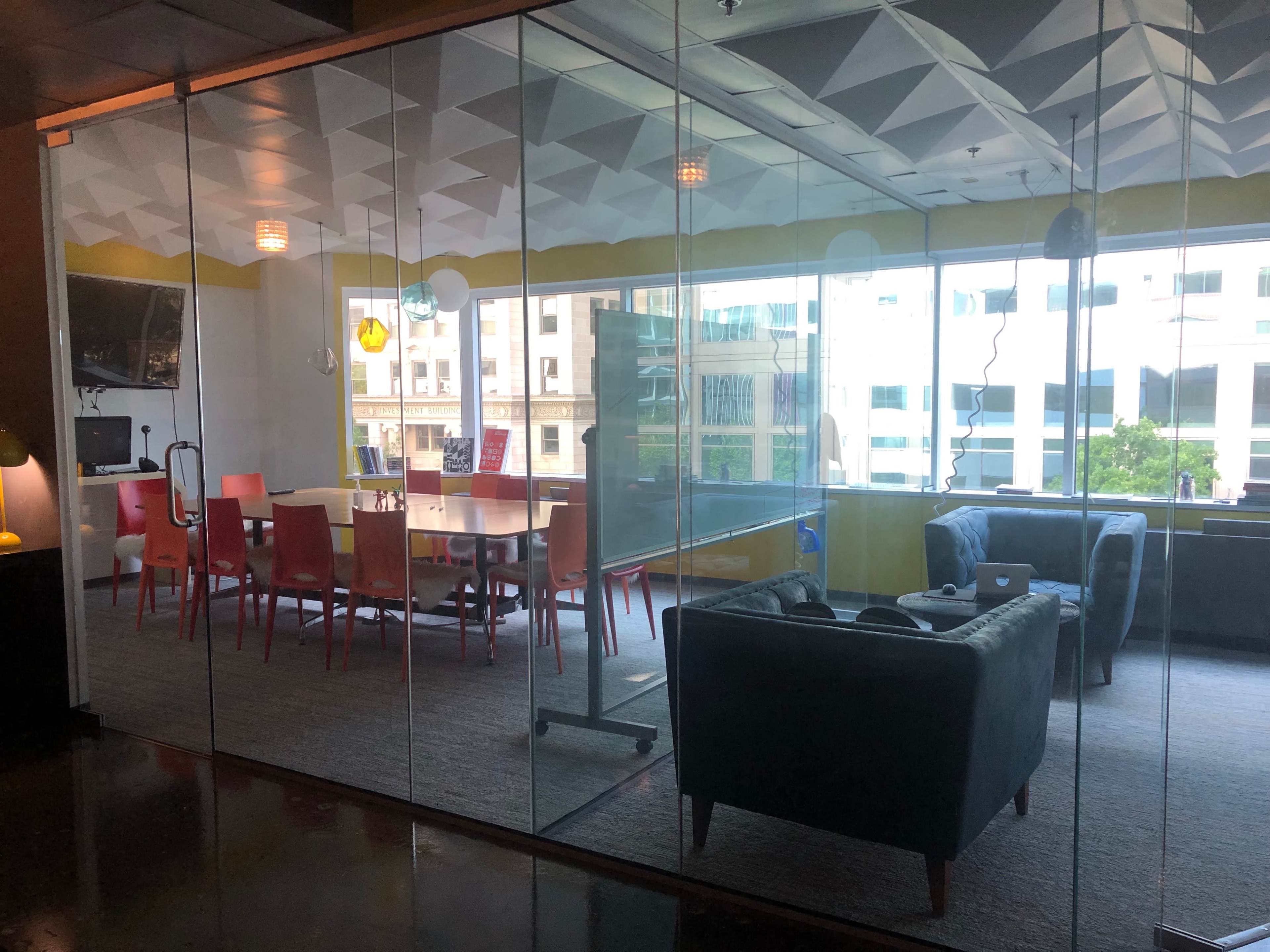 A modern conference room with a glass wall, featuring colorful chairs around a large table, and a lounge area with a couch and workspace.