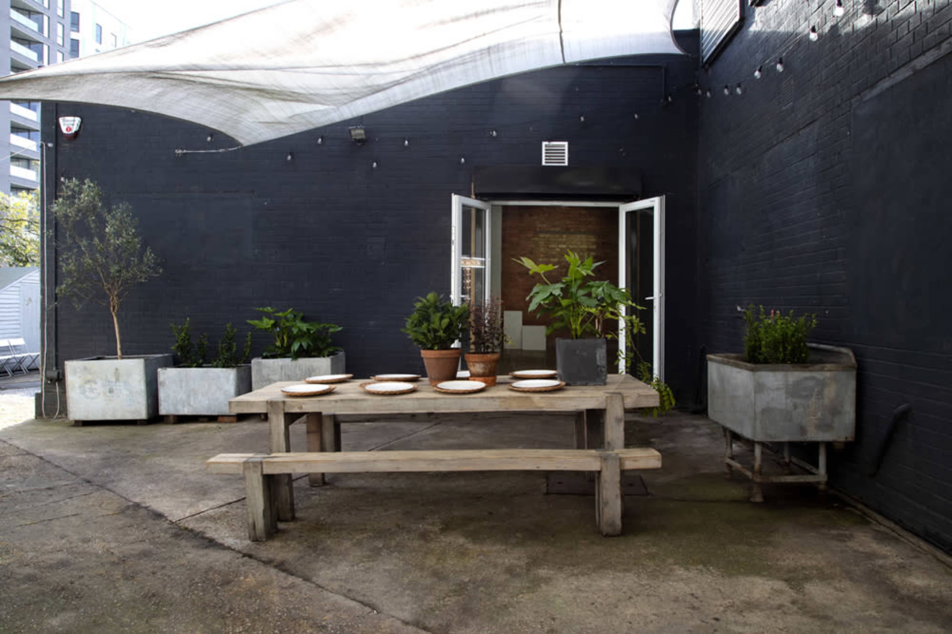 A wooden table is set with plates in a shaded outdoor area surrounded by potted plants and a dark brick wall.