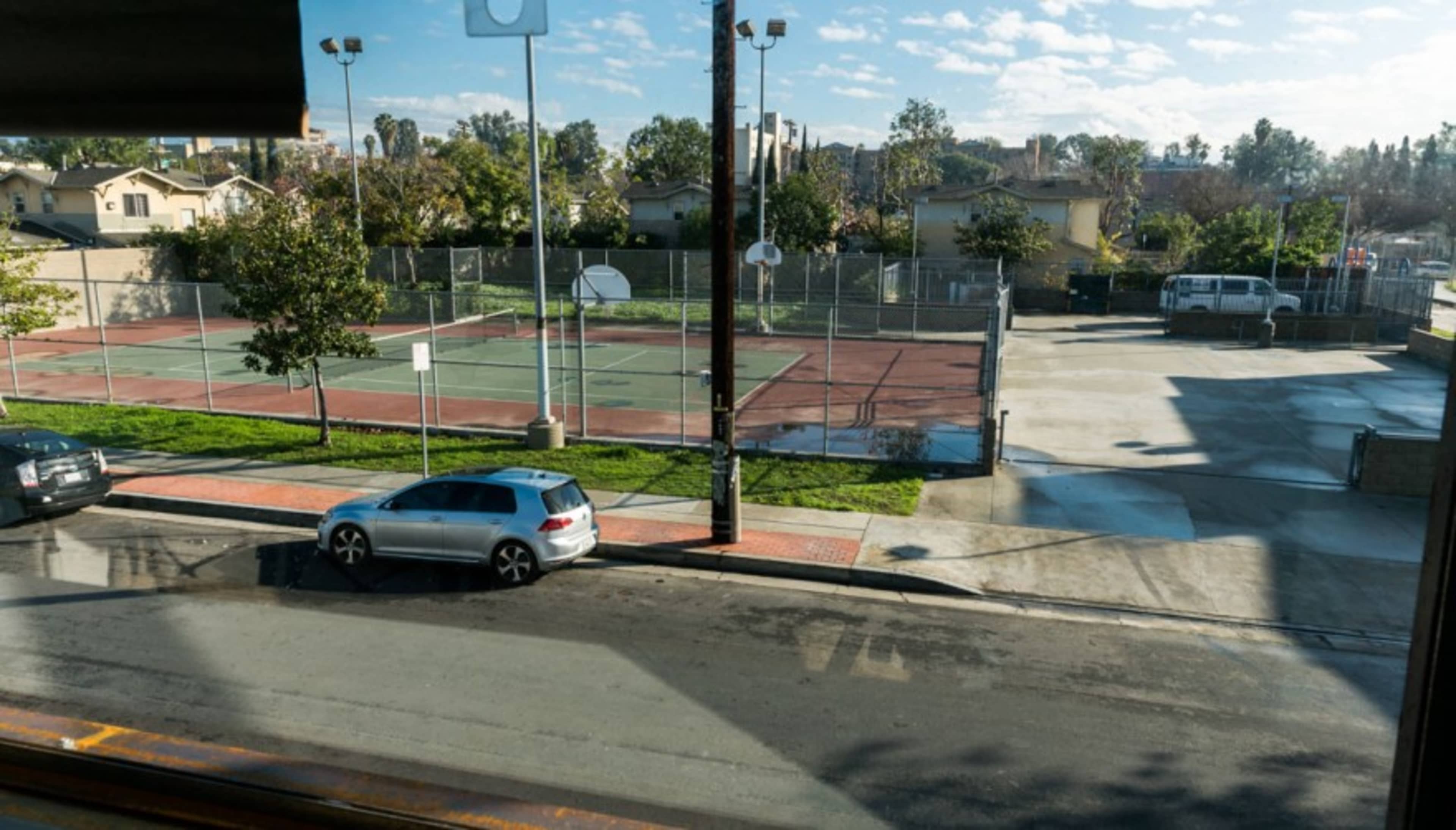 The image shows an outdoor tennis court area bordered by residential buildings, with a street and parked cars visible in the foreground.