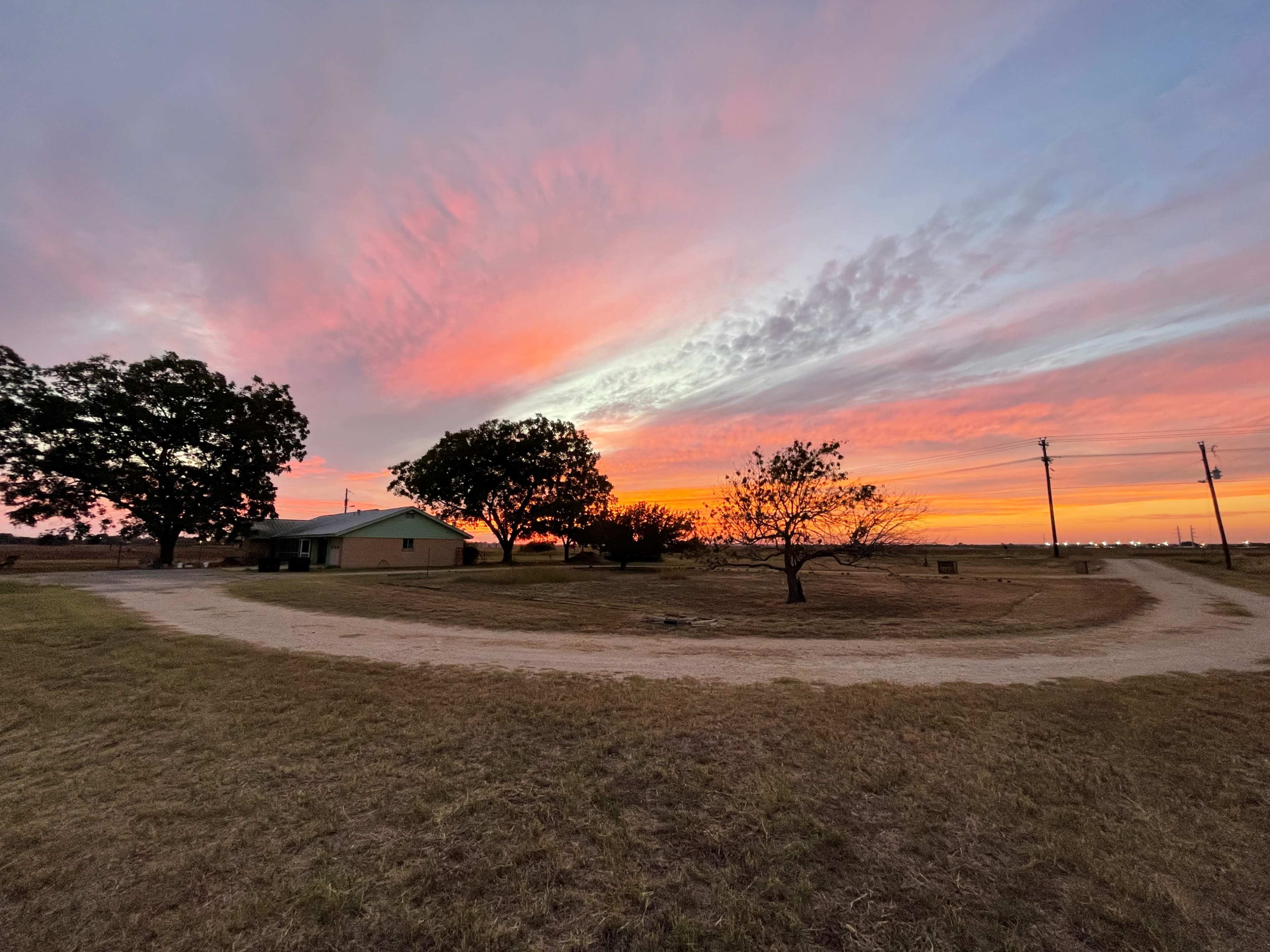 A winding dirt road leads to a house under a colorful sunset, with trees silhouetted against the sky.
