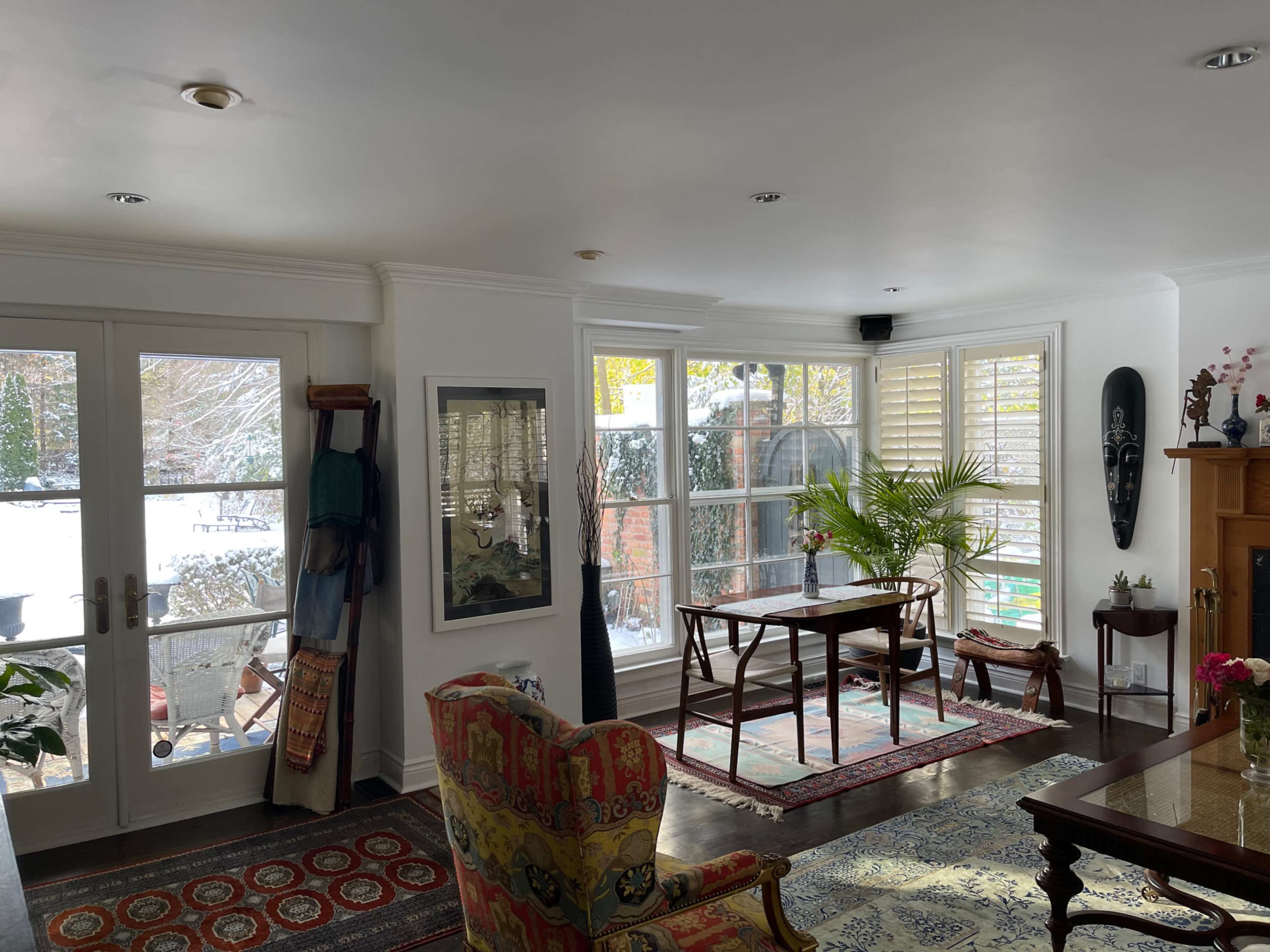 A spacious living room with large windows letting in natural light, a wooden table in the center, and various furnishings including chairs and a rug.